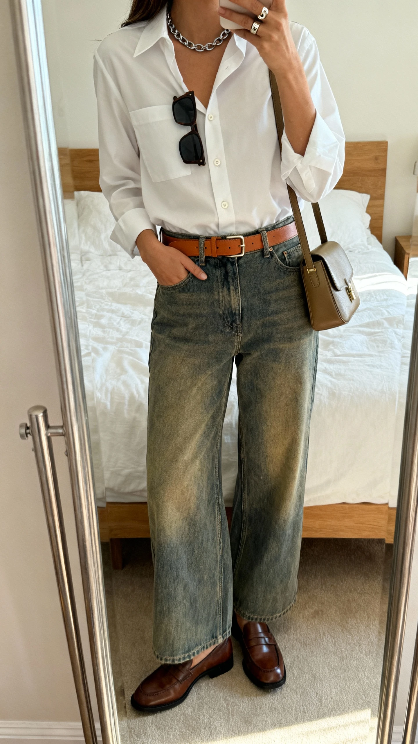 iPhone mirror selfie of a woman in high-waisted wide-leg vintage-wash jeans, crisp white tailored button-down, leather loafers, tan leather belt, medium-structured crossbody, simple chain necklace, signet ring, rectangular sunglasses tucked in shirt; face not visible, cropped at shoulders, bedroom mirror, morning light, iPhone photo quality.