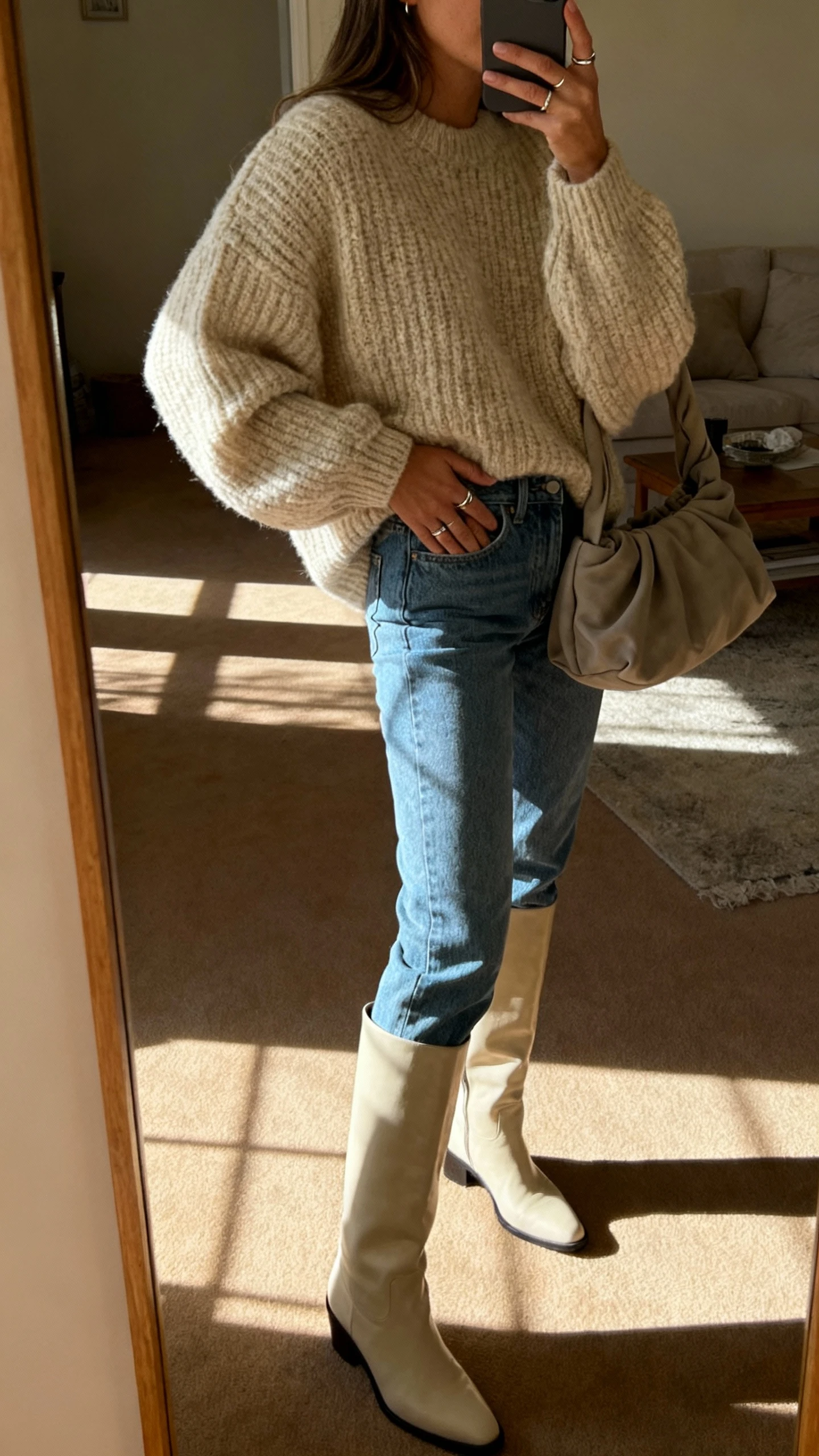 iPhone mirror selfie of a woman in mom jeans, a cozy oversized chunky knit sweater, statement knee-high boots, minimal rings, and a slouchy tote, face not visible, side angle in a living room mirror with morning light, iPhone photo quality.