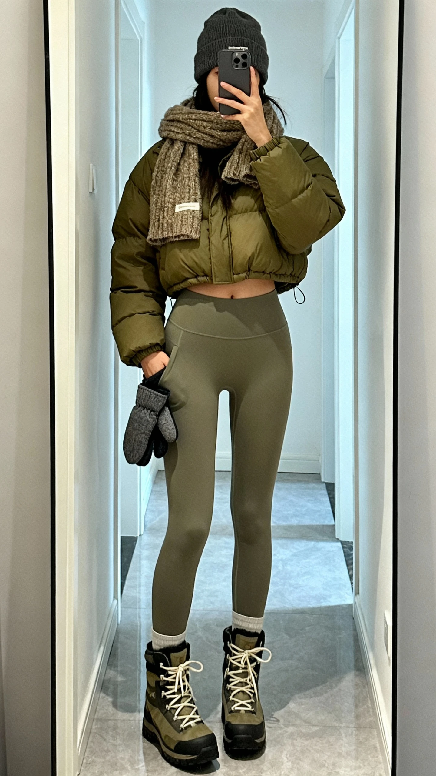 iPhone mirror selfie of a woman in thermal leggings, cropped puffer jacket in olive, chunky scarf, beanie, lace-up winter boots, mittens tucked in pocket, face not visible, hallway full-length mirror with cool daylight, iPhone photo quality