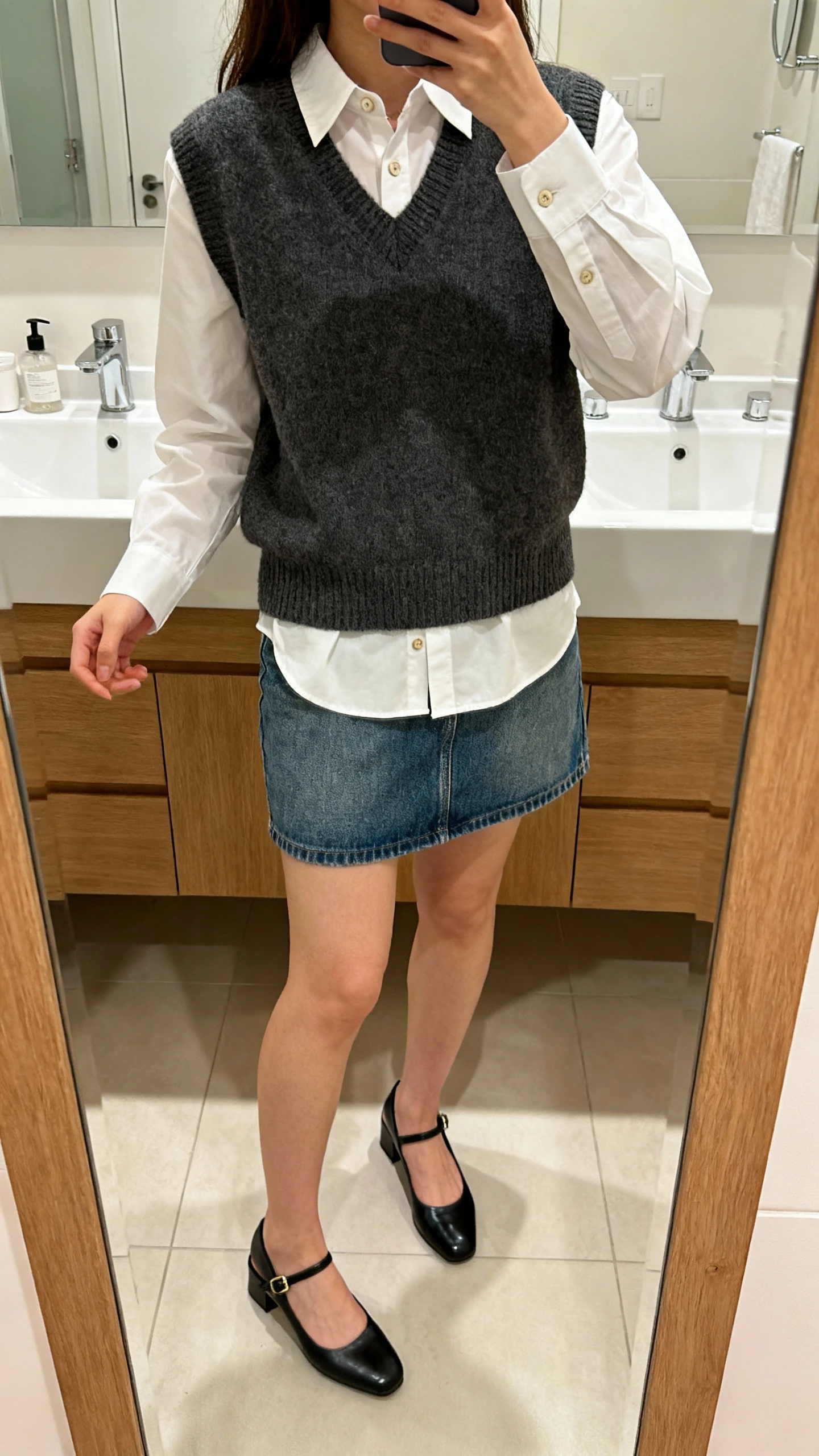 iPhone mirror selfie of a woman layered in a crisp button-up under a sweater vest with a denim mini skirt and slingback shoes, face not visible, cropped at neck, bathroom mirror, indoor lighting, iPhone photo quality.