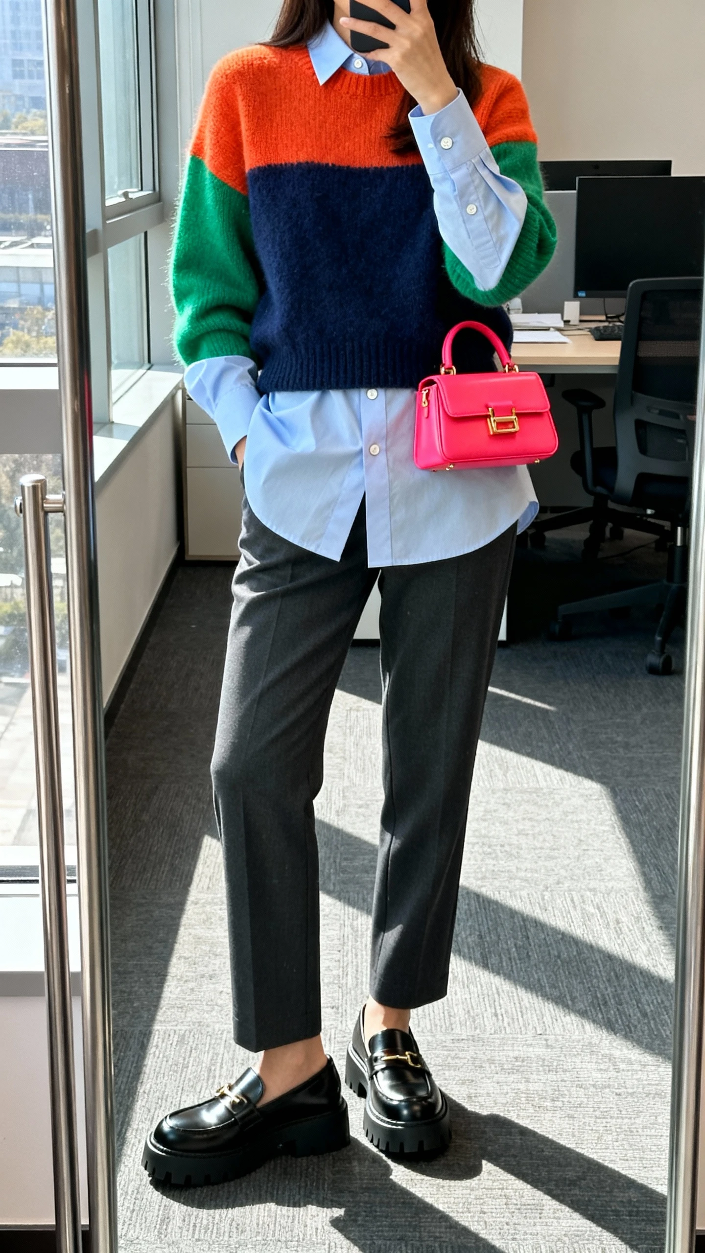 iPhone mirror selfie of a woman layering a color-pop sweater over a crisp button-down, straight trousers, chunky loafers, bright mini bag, face not visible, cropped at shoulders, office mirror, natural window light, iPhone photo quality.