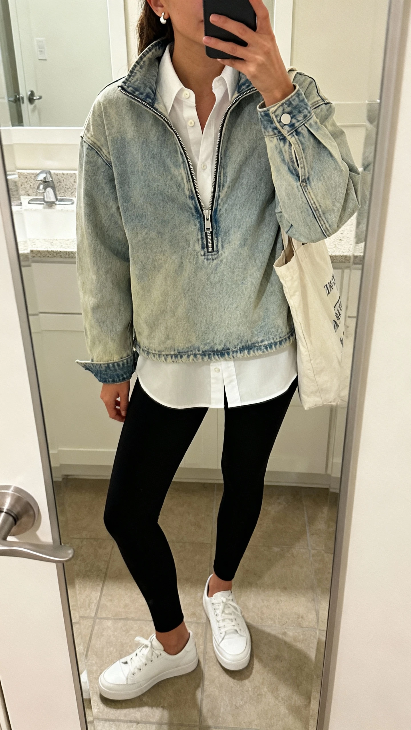 iPhone mirror selfie of a woman showing denim jacket + button-up layering hack: white button-up under light-wash denim jacket half-zipped, black leggings, white low-top sneakers, tote bag, simple studs, face not visible, bathroom mirror, soft indoor lighting, iPhone photo quality