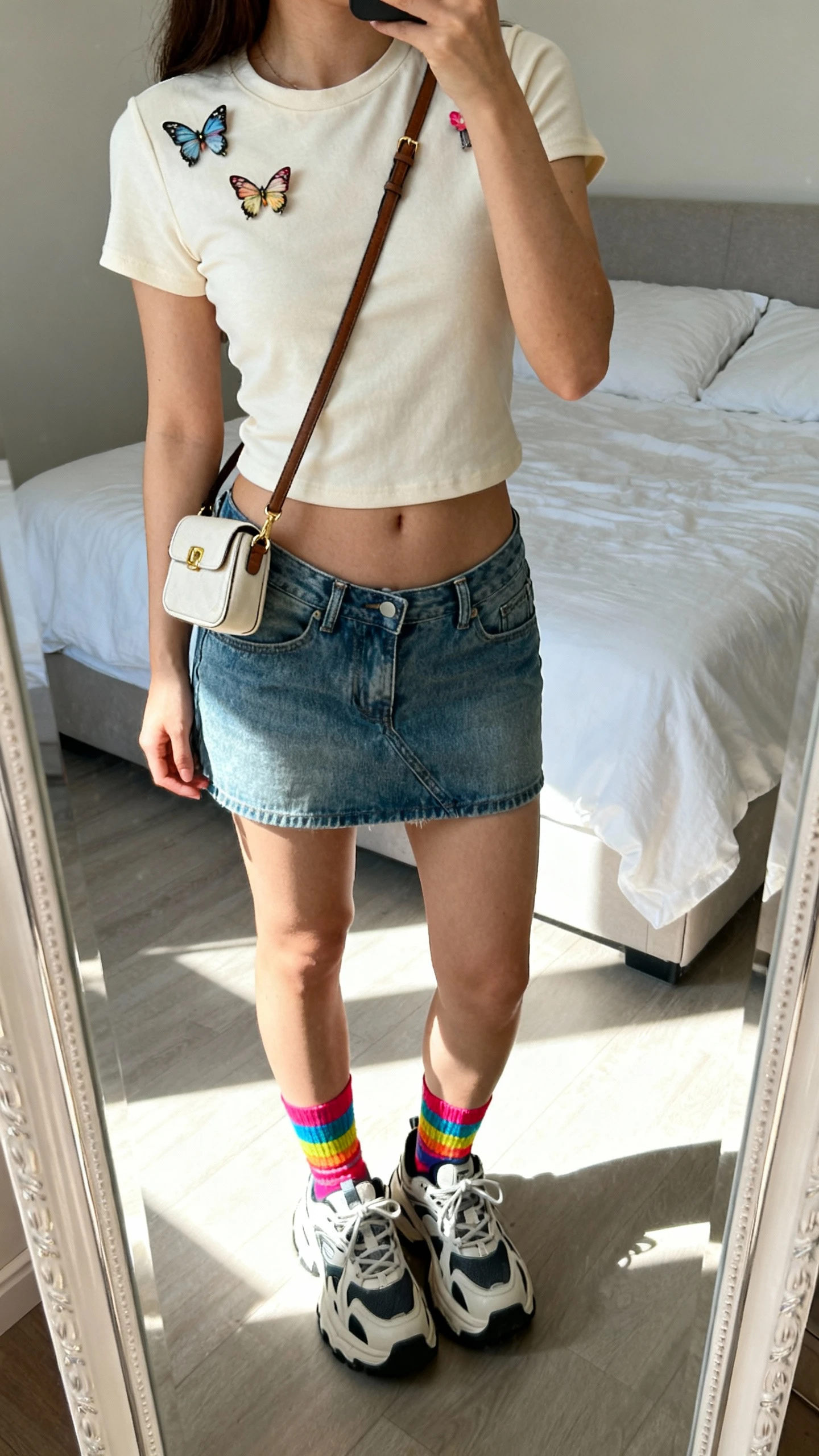 iPhone mirror selfie of a woman wearing a baby tee, low-rise denim mini skirt, chunky sneakers, colorful socks, tiny shoulder bag, and butterfly clips, face not visible, cropped at shoulders, bedroom mirror with bright daylight, iPhone photo quality.