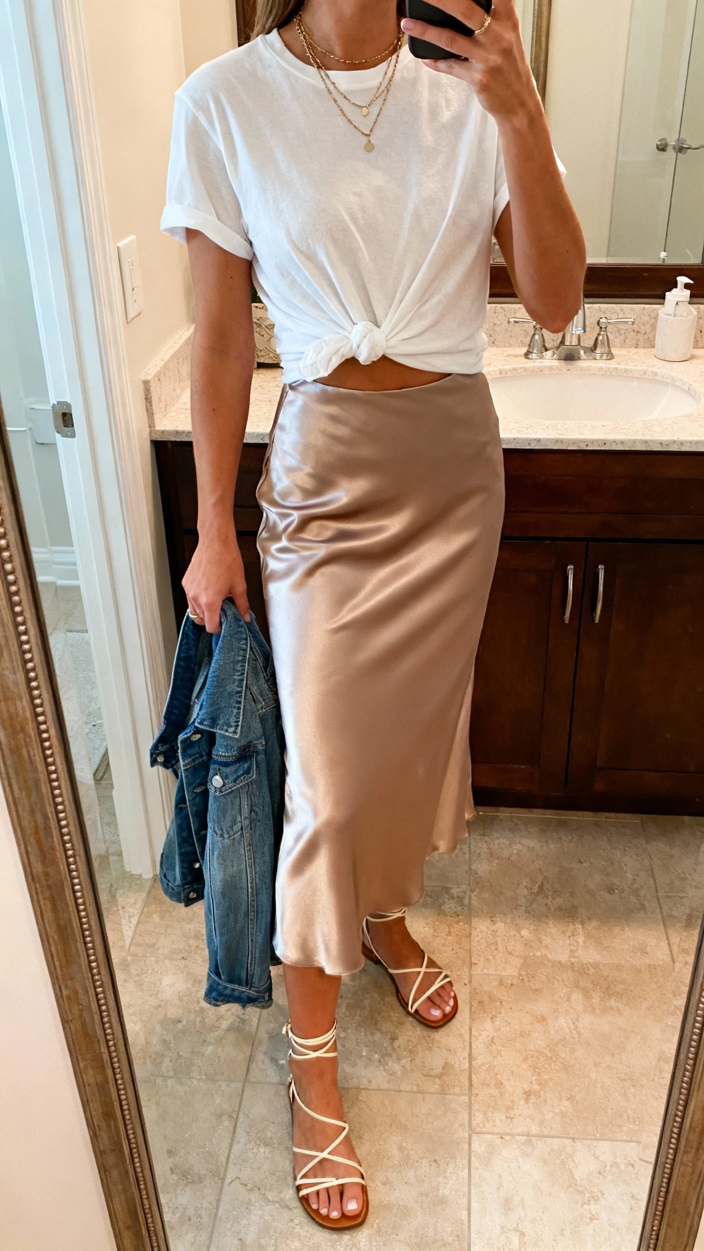 iPhone mirror selfie of a woman wearing a bias-cut satin midi skirt with a relaxed white tee knotted at the waist, delicate layered necklaces, denim jacket in hand, and strappy sandals, face not visible, bathroom mirror, warm indoor lighting, iPhone photo quality.