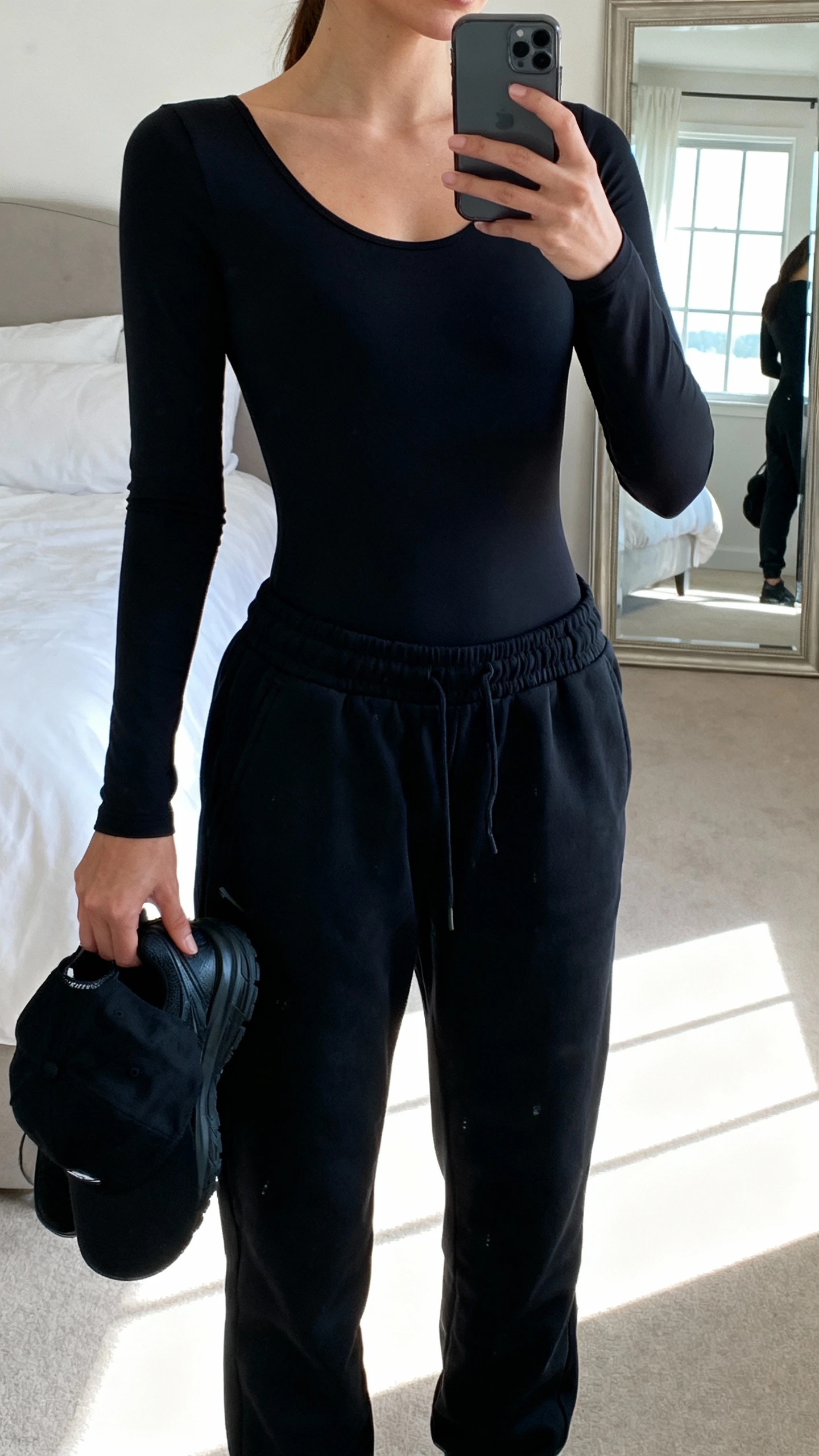 iPhone mirror selfie of a woman wearing a black bodysuit and black training joggers, face not visible, cropped at neck, full-length mirror in a minimalist bedroom with window light, black running shoes and a subtle black baseball cap in hand, iPhone photo quality