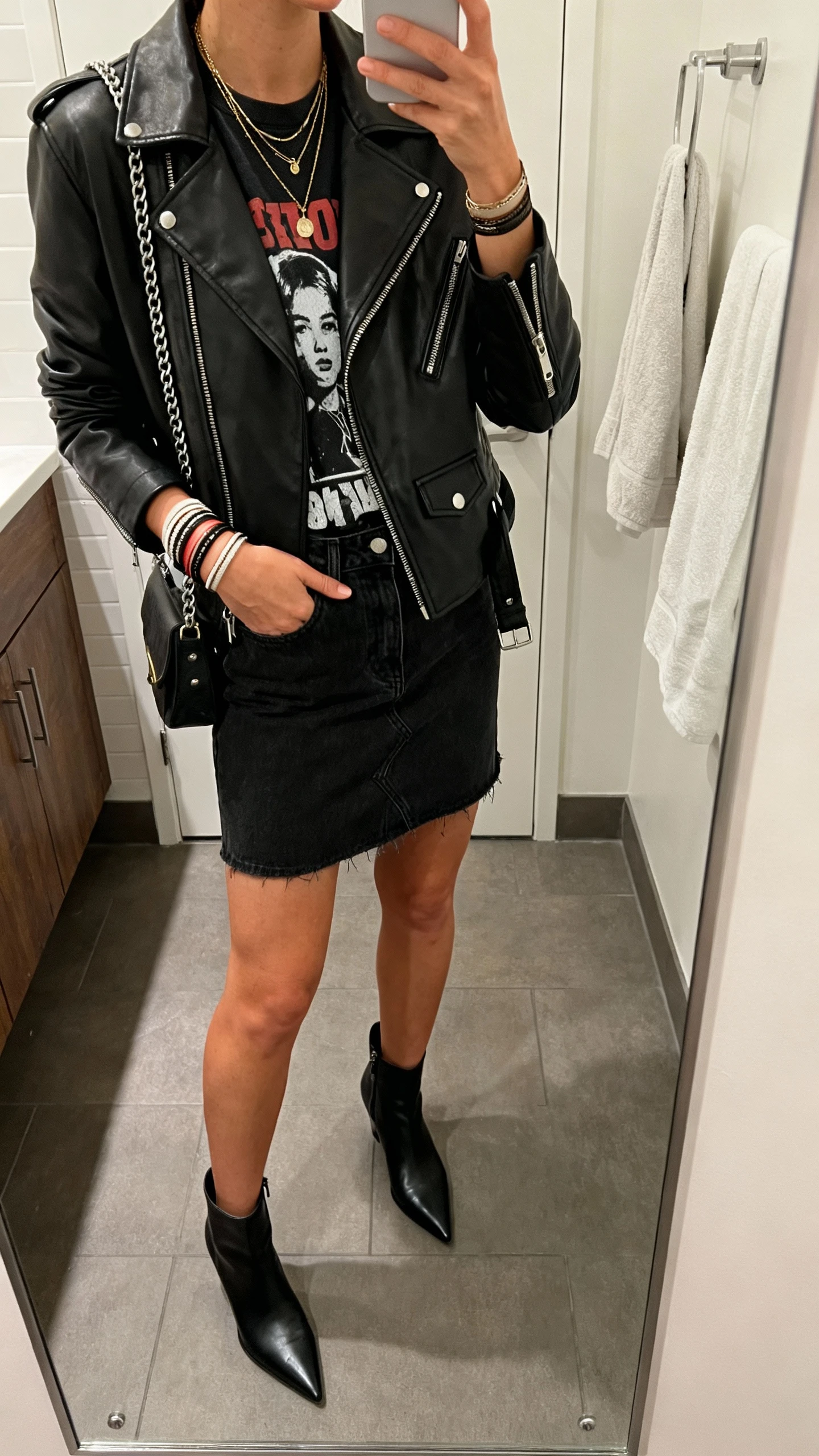 iPhone mirror selfie of a woman wearing a black denim mini skirt, black leather moto jacket over a graphic tee, pointed ankle boots, a chain-strap crossbody, layered necklaces, and stacked cuffs; face not visible, cropped at shoulders, bathroom mirror, moody indoor lighting, iPhone photo quality.