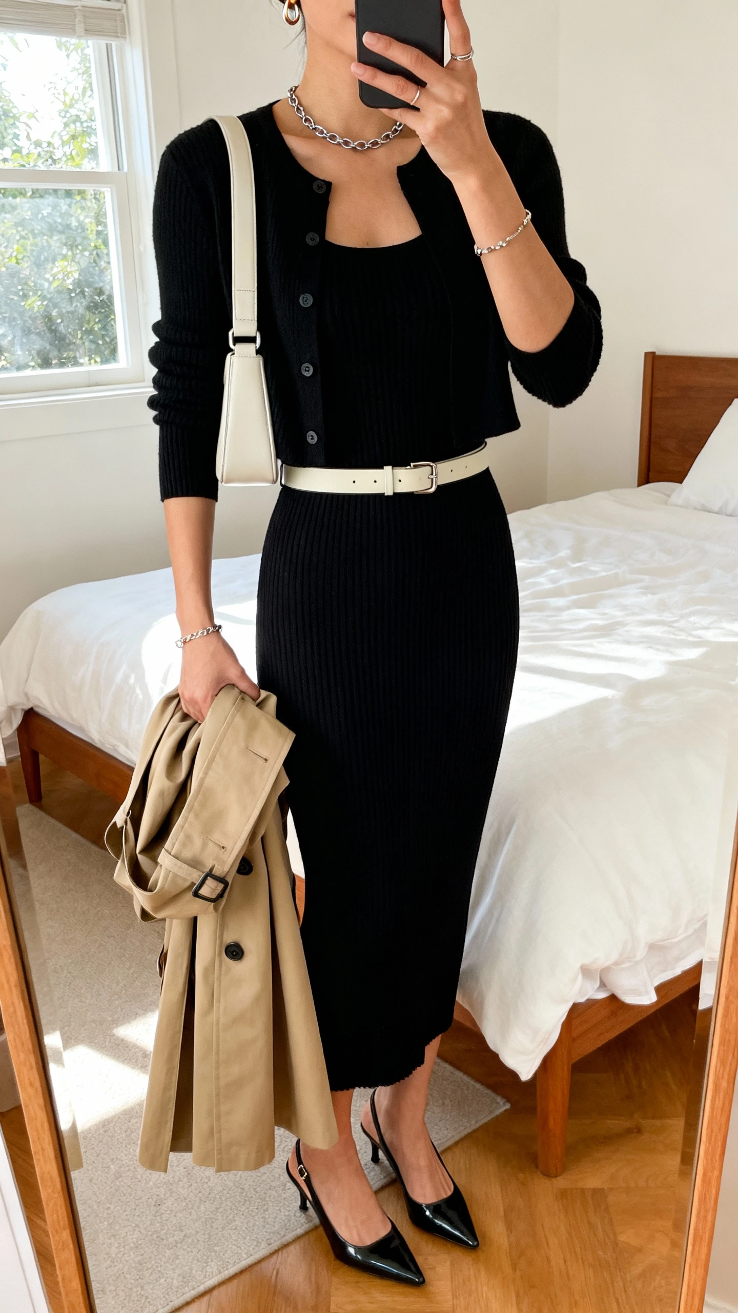 iPhone mirror selfie of a woman wearing a black knit column dress, cropped cardigan, sleek slingback heels, clean-lined shoulder bag, delicate chain, bracelet, small hoops, and a light waist belt, with a trench draped on forearm; face not visible, cropped at neck, bedroom mirror, afternoon window light, iPhone photo quality.