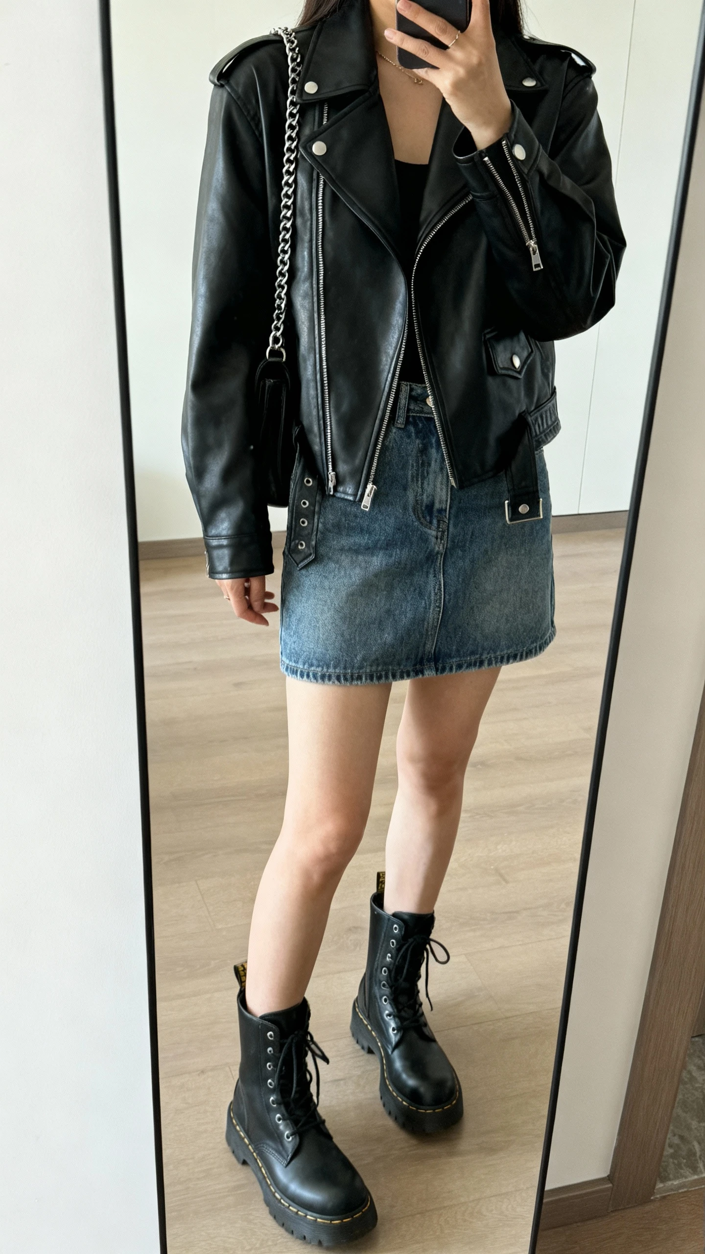 iPhone mirror selfie of a woman wearing a black leather moto jacket over a denim mini skirt with combat boots and a chain-strap bag, face not visible, cropped at shoulders, full-length mirror, cool indoor lighting, iPhone photo quality.