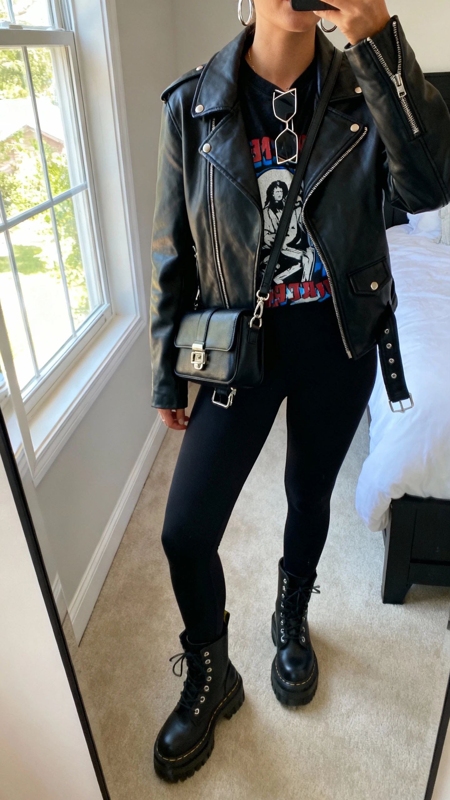 iPhone mirror selfie of a woman wearing a black leather moto jacket, graphic tee, matte black leggings, chunky combat boots, black crossbody with metal hardware, angular sunglasses and silver hoops, face not visible, cropped at shoulders, bedroom mirror, natural window light, iPhone photo quality.
