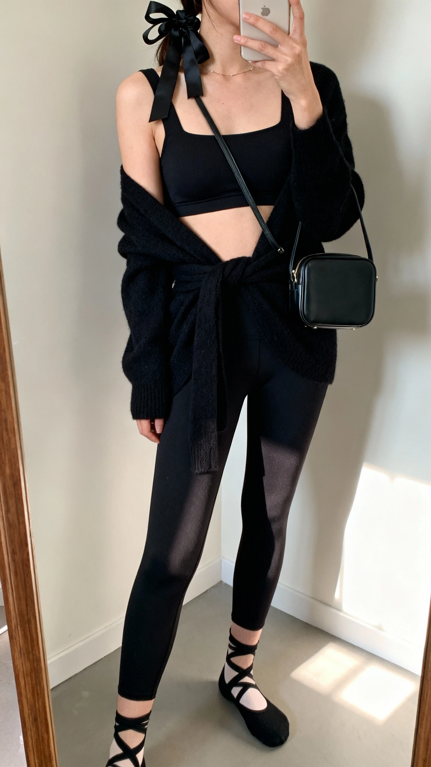 iPhone mirror selfie of a woman wearing a black longline bra with a square neckline, black stirrup leggings, black wrap cardigan, grip socks, a delicate black ribbon scrunchie in hair, and a compact black shoulder bag, face not visible, studio corner mirror, soft morning light, iPhone photo quality.