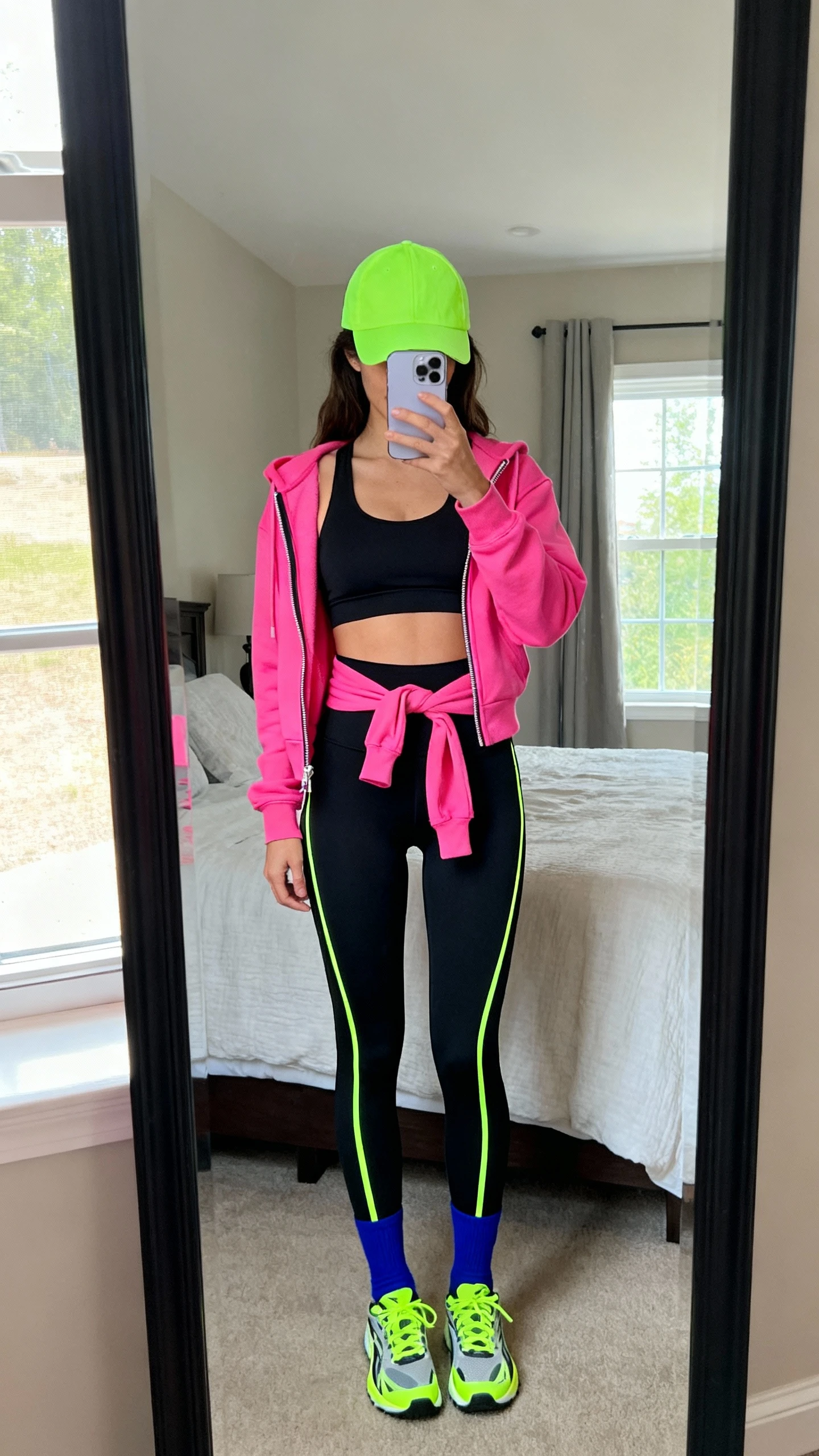 iPhone mirror selfie of a woman wearing a black sports bra and black high-waist leggings with electric neon accents: hot-pink zip-up hoodie tied at waist, lime-green baseball cap, cobalt blue crew socks, and neon-accent trainers, face not visible, cropped at neck, bedroom mirror, natural window light, iPhone photo quality.