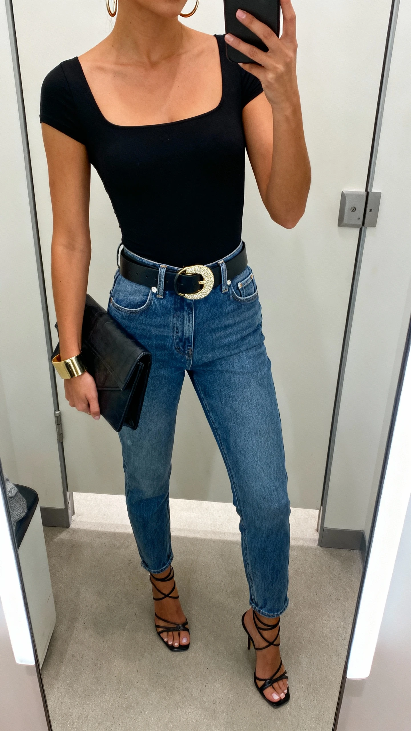 iPhone mirror selfie of a woman wearing a black square-neck stretch bodysuit tucked into high-rise mom jeans with belt loops, a statement belt with bold buckle, strappy heels, hoop earrings, a cuff bracelet, and a structured clutch, face not visible, cropped at shoulders, dressing-room mirror, indoor lighting, iPhone photo quality.