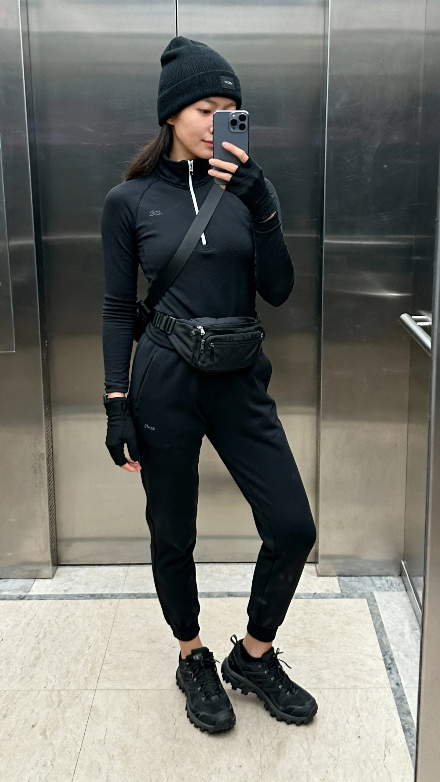 iPhone mirror selfie of a woman wearing a black thermal long-sleeve base layer, black joggers with tapered ankles, black half-zip fleece, black trail-inspired sneakers, black beanie, crossbody belt bag, and thin black gloves, face not visible, elevator mirror, cool indoor lighting, iPhone photo quality.