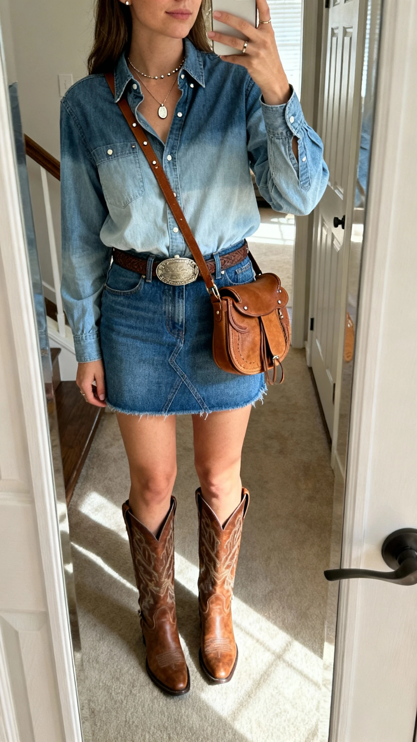 iPhone mirror selfie of a woman wearing a blue denim mini skirt with a different-wash chambray shirt tucked in, mid-calf Western boots, a belt with a Western buckle, tan saddle crossbody, a simple pendant necklace, and studs; face not visible, cropped at nose, entryway full-length mirror, natural daylight, iPhone photo quality.