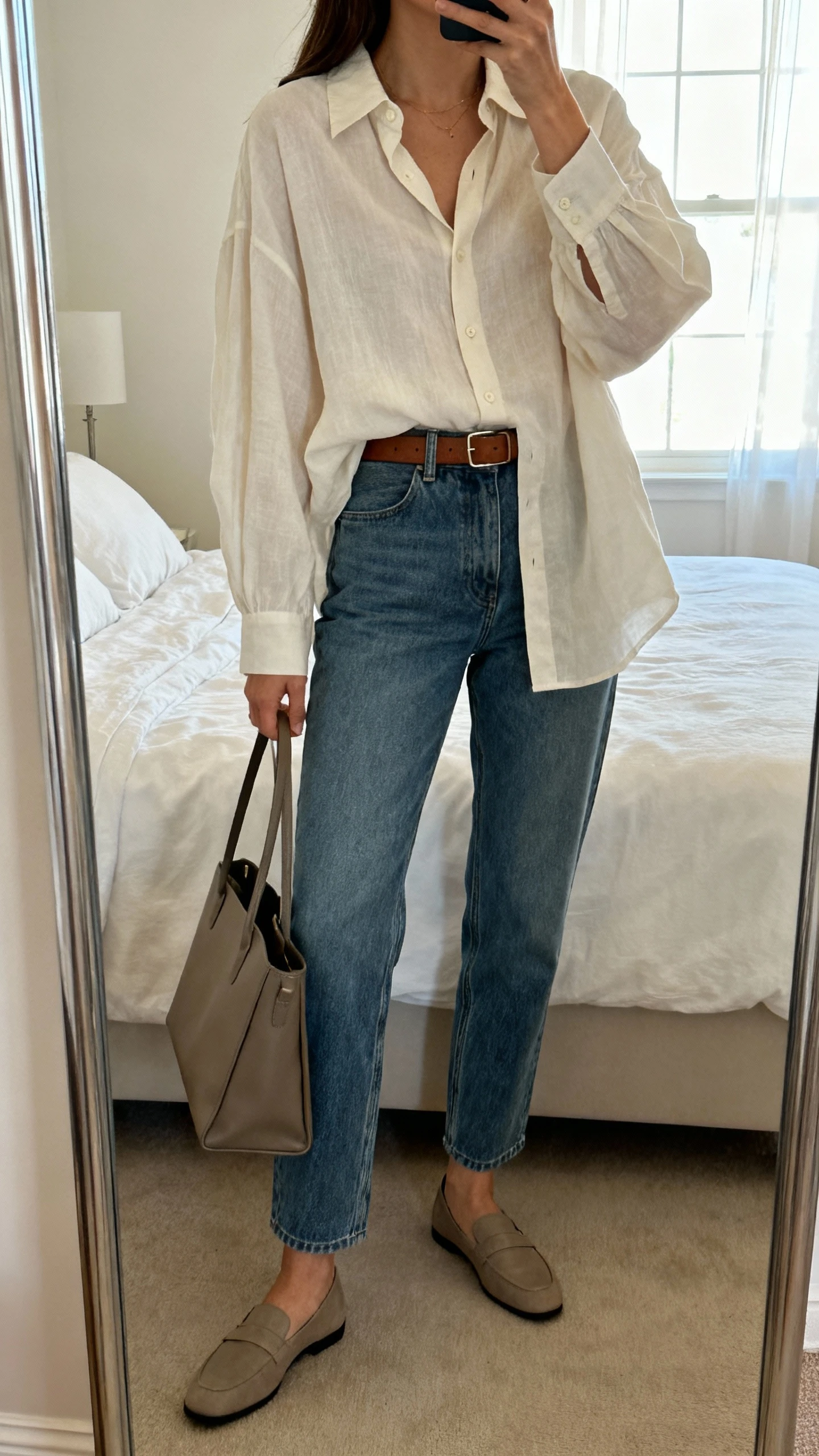 iPhone mirror selfie of a woman wearing a breezy oversized button-down shirt tucked into straight-leg jeans with a slim leather belt, minimalist loafers, and a structured tote, face not visible, cropped at neck, bedroom mirror, morning window light, iPhone photo quality.