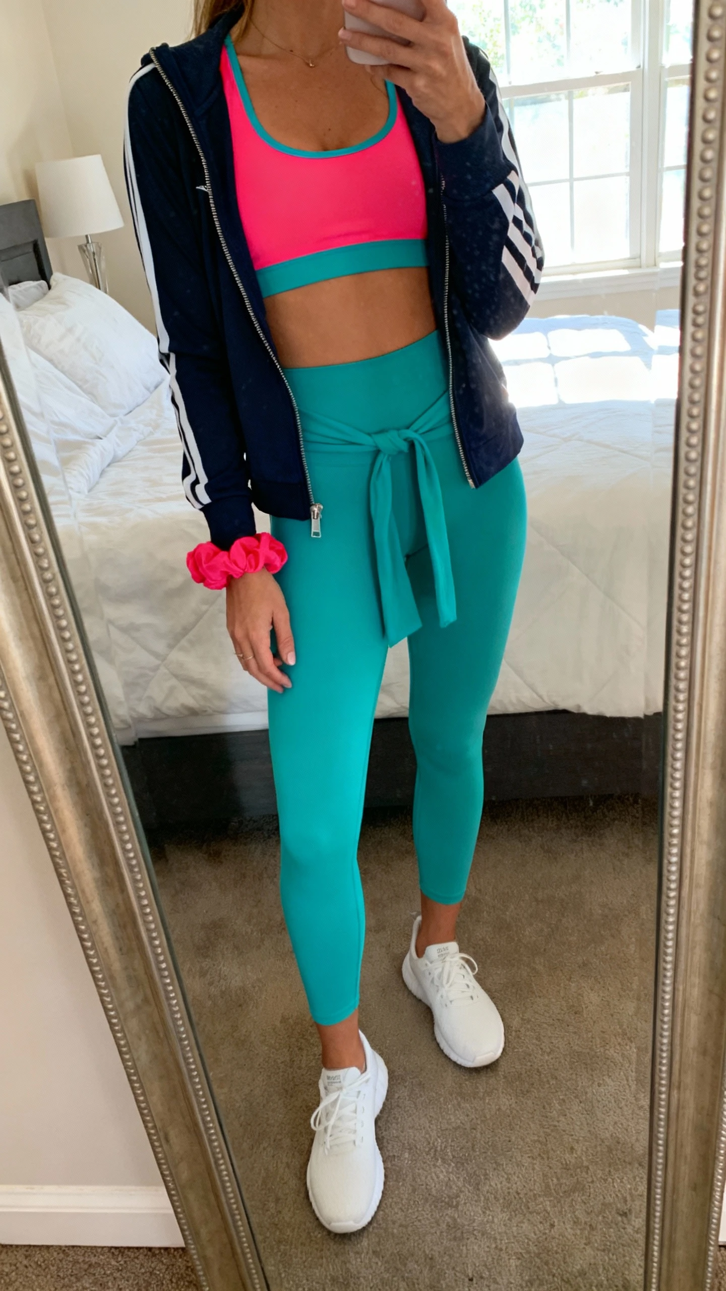 iPhone mirror selfie of a woman wearing a bright color-pop matching sports bra and high-waist leggings set with white cushioned trainers, slim zip-up track jacket tied at waist, and a bold scrunchie on her wrist, face not visible, cropped at neck, bedroom mirror, morning window light, iPhone photo quality