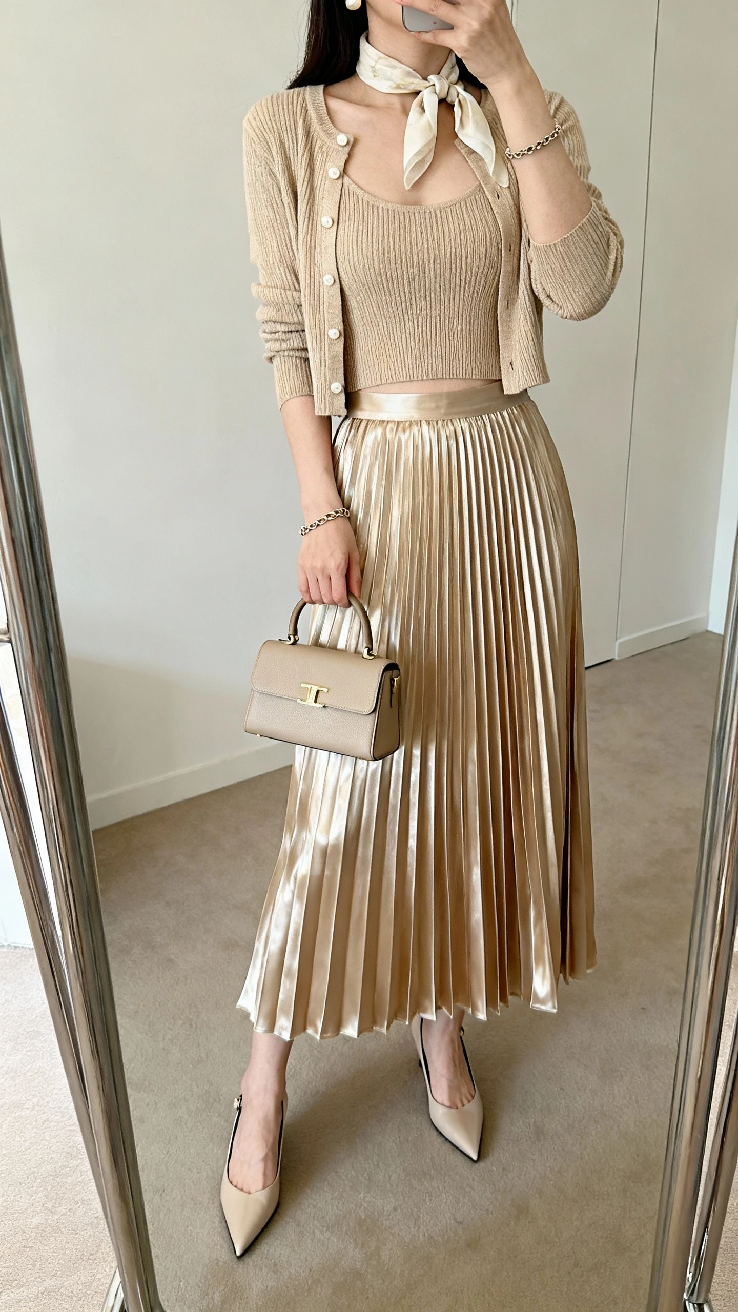 iPhone mirror selfie of a woman wearing a champagne silky pleated midi skirt with matching knit camisole and cropped cardigan, pointed-toe slingbacks, structured mini top-handle bag, pearl studs, thin chain bracelet, light neckerchief; face not visible, cropped at neck, full-length mirror, soft indoor lighting, iPhone photo quality.