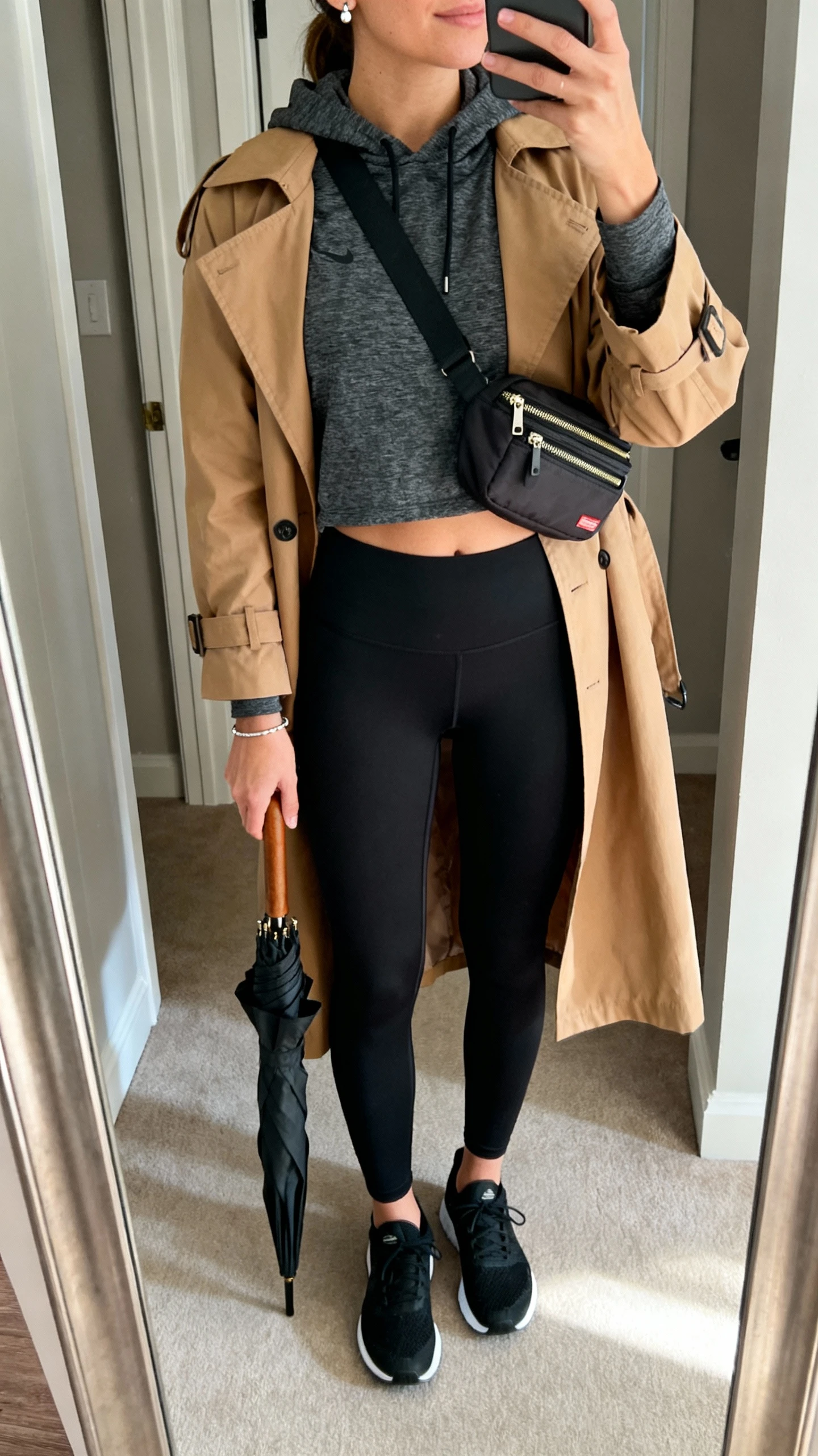 iPhone mirror selfie of a woman wearing a charcoal moisture-wicking hoodie, black high-waisted sculpt leggings, a camel lightweight trench coat, black running shoes, a crossbody bag with zip compartments, simple stud earrings, a minimal bracelet, and a foldable umbrella tucked under arm; face not visible, cropped at neck, entryway mirror, indoor daylight, iPhone photo quality.