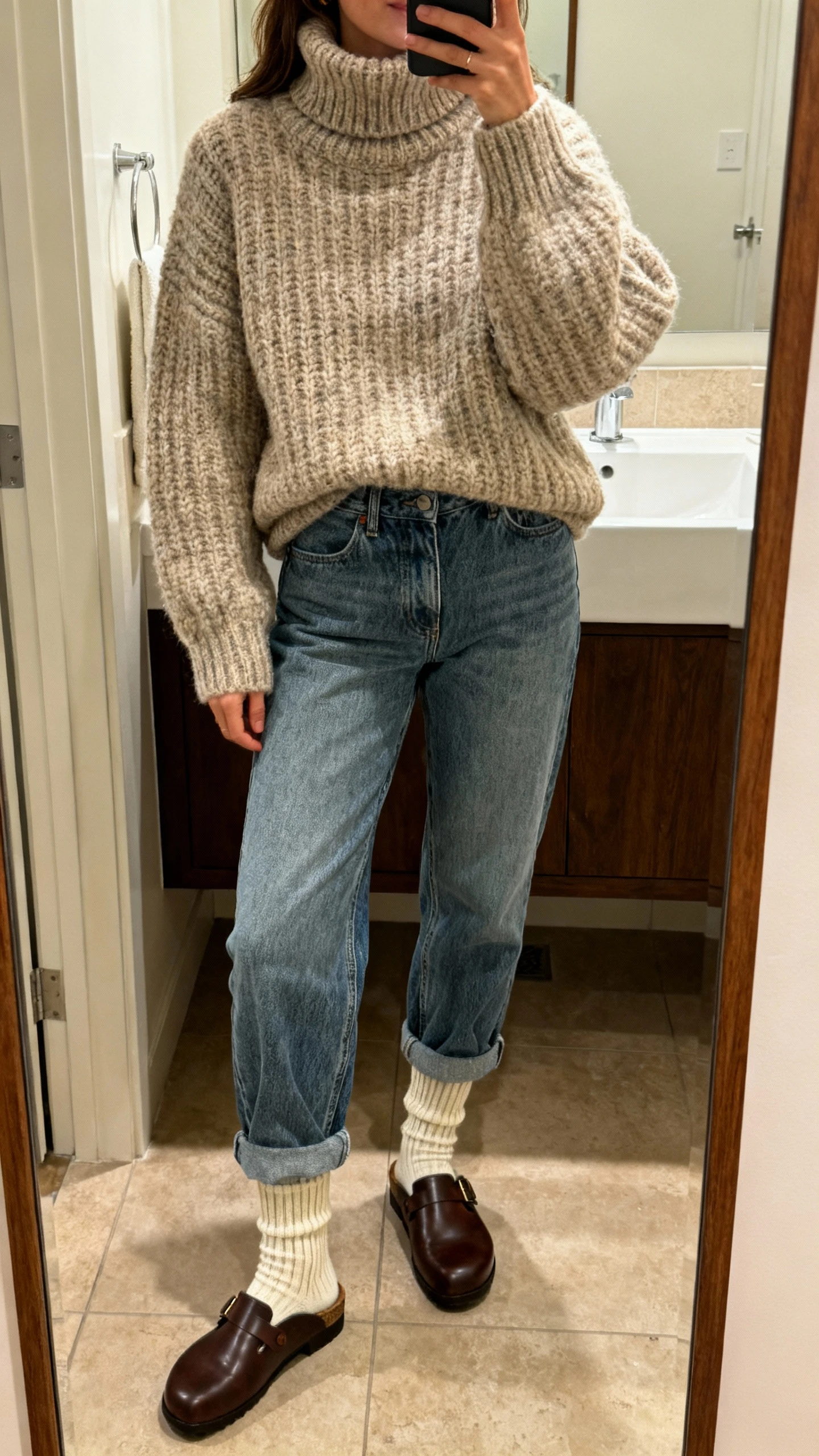 iPhone mirror selfie of a woman wearing a chunky cozy turtleneck sweater with relaxed mom jeans, ribbed socks and classic leather clogs, face not visible, cropped at shoulders, bathroom mirror, warm indoor light, iPhone photo quality.
