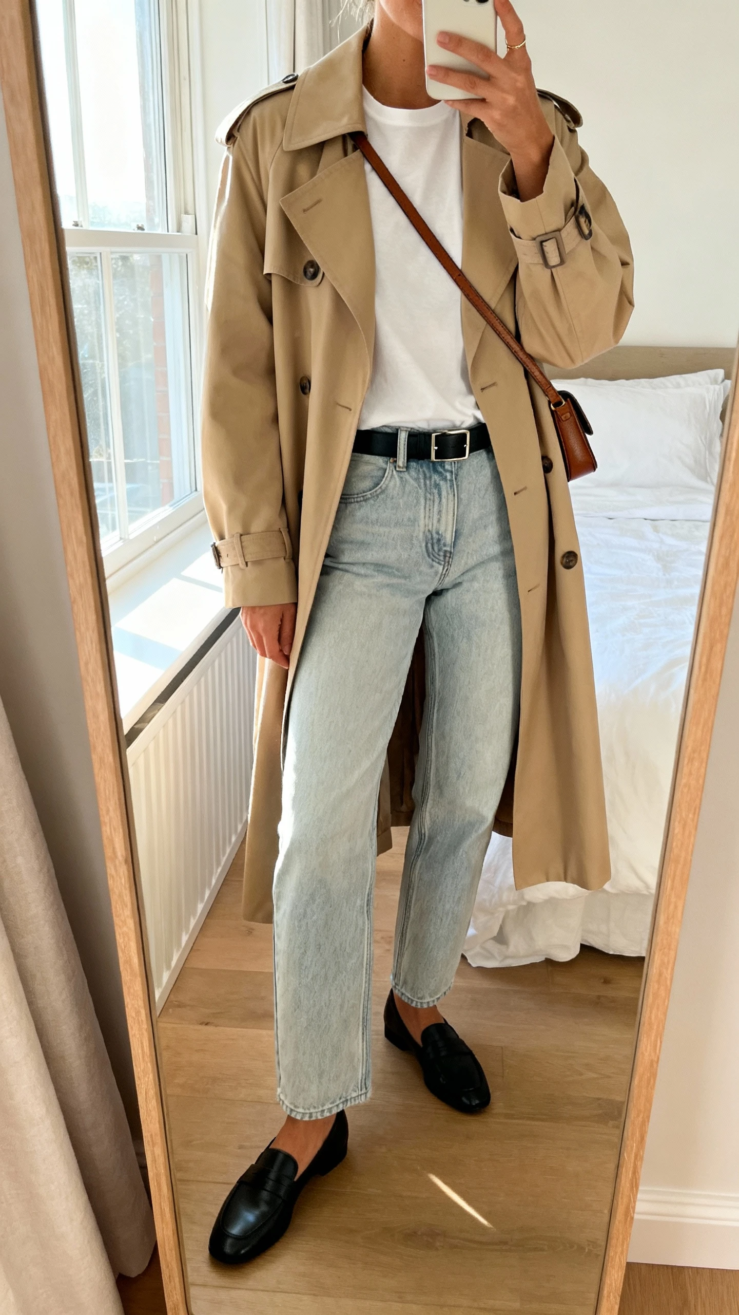 iPhone mirror selfie of a woman wearing a classic beige trench coat over light-wash straight-leg denim, white tee, black loafers, slim black belt, and a simple leather crossbody, face not visible, cropped at neck, bedroom mirror with morning window light, iPhone photo quality.