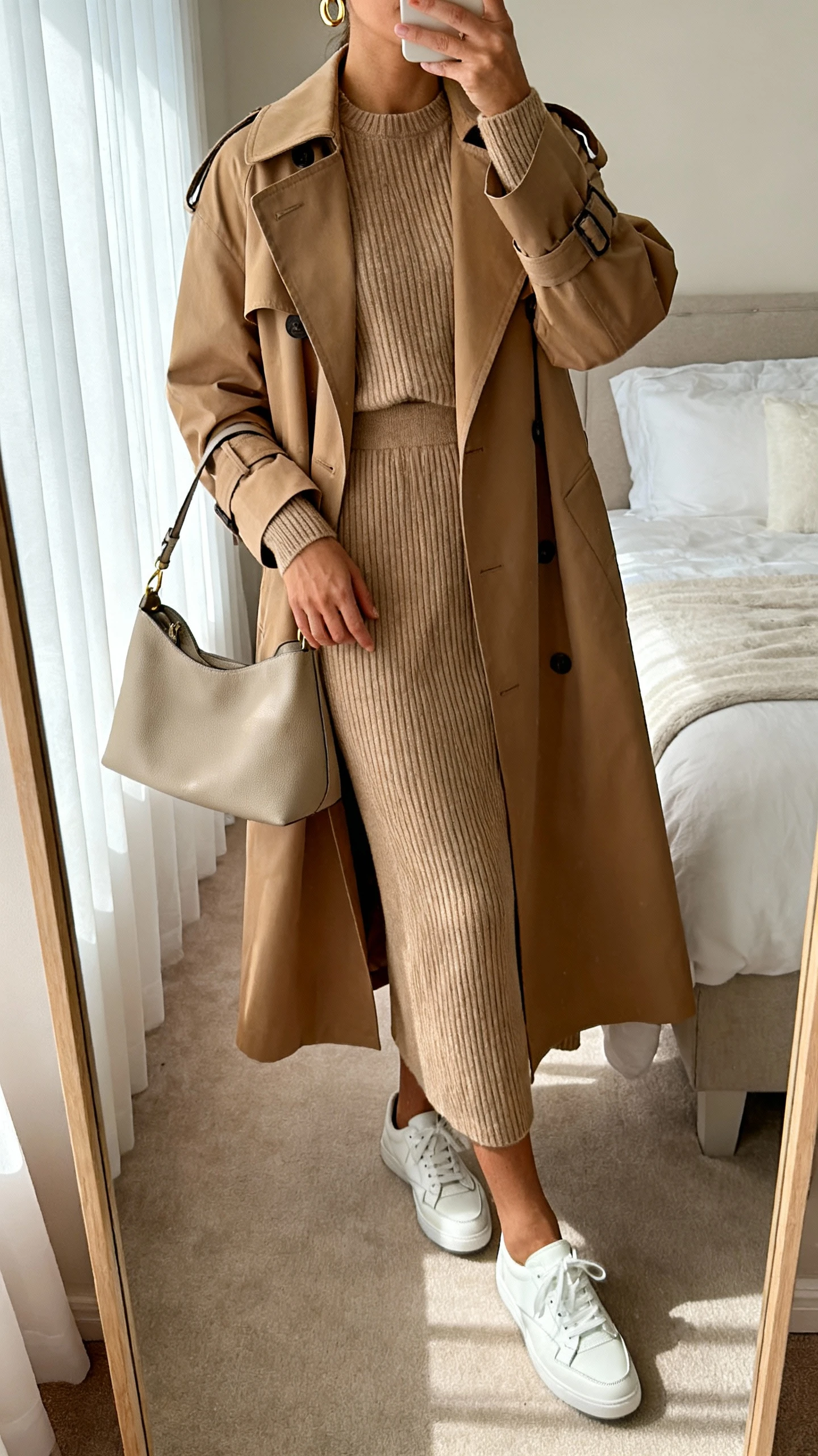 iPhone mirror selfie of a woman wearing a classic camel trench coat over a matching ribbed knit set (crewneck sweater and midi skirt), white leather sneakers, structured tote, delicate gold hoops, face not visible, cropped at neck, bedroom mirror, morning window light, iPhone photo quality.