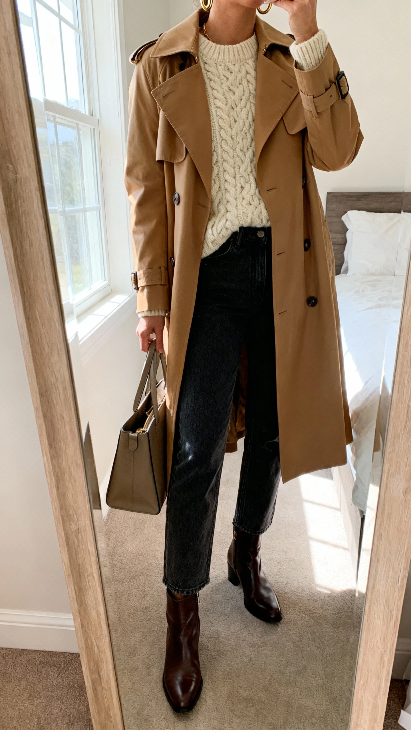 iPhone mirror selfie of a woman wearing a classic camel trench coat layered over a chunky cream knit sweater, straight-leg dark jeans, sleek leather ankle boots, minimal gold hoops, and a structured tote, face not visible, cropped at neck, bedroom mirror, natural window light, iPhone photo quality.