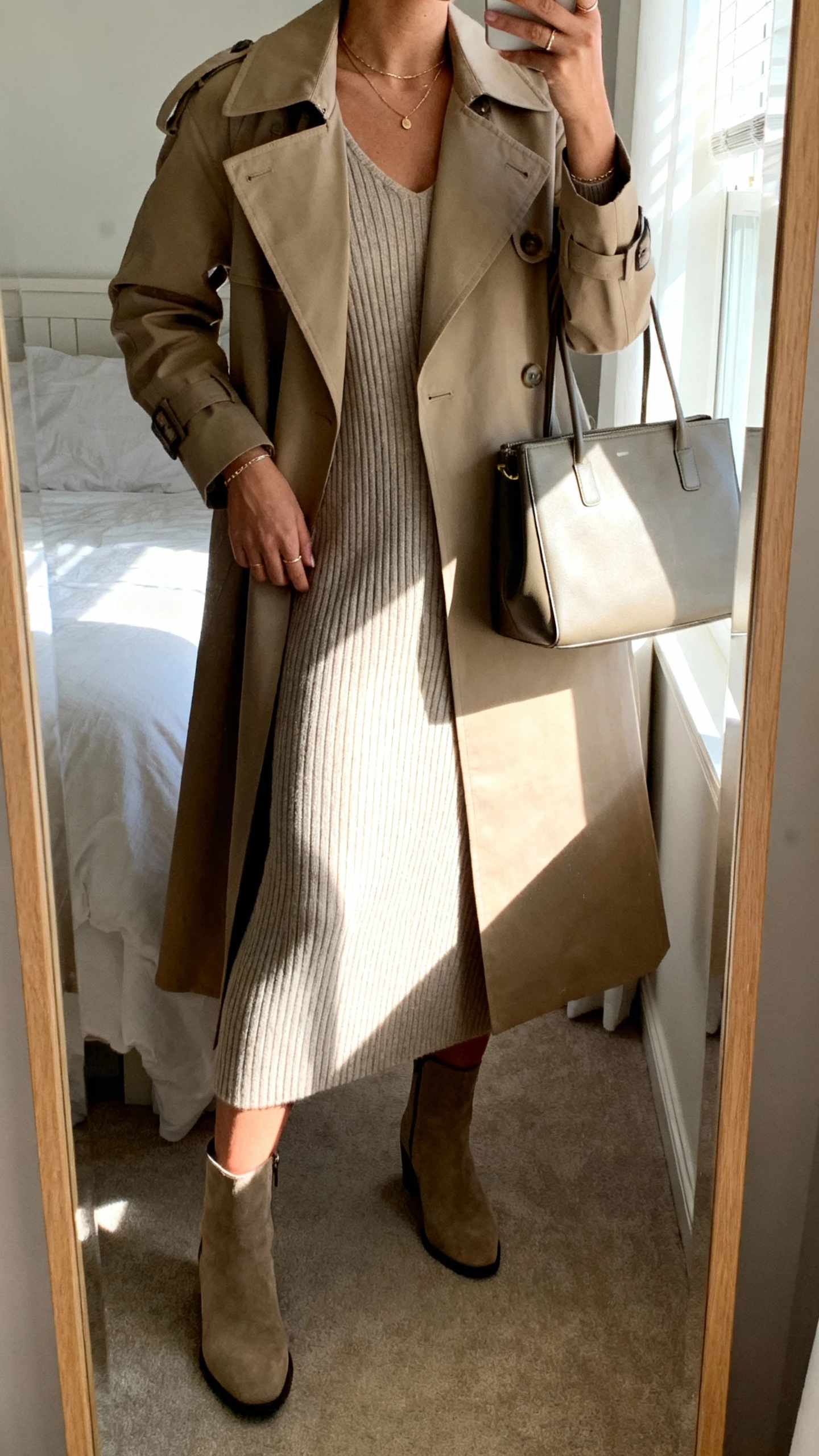iPhone mirror selfie of a woman wearing a classic trench coat over a ribbed knit midi dress, ankle boots, minimal jewelry, and a structured tote, face not visible, cropped at neck, bedroom mirror with morning window light, iPhone photo quality.