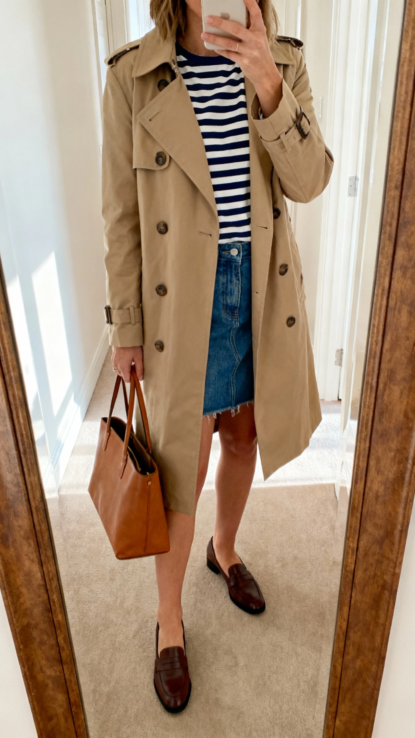 iPhone mirror selfie of a woman wearing a classic trench coat over a navy-and-cream Breton striped tee, denim mini skirt, low-heel loafers, and a simple leather tote, face not visible, cropped at shoulders, hallway mirror with soft natural light, iPhone photo quality.