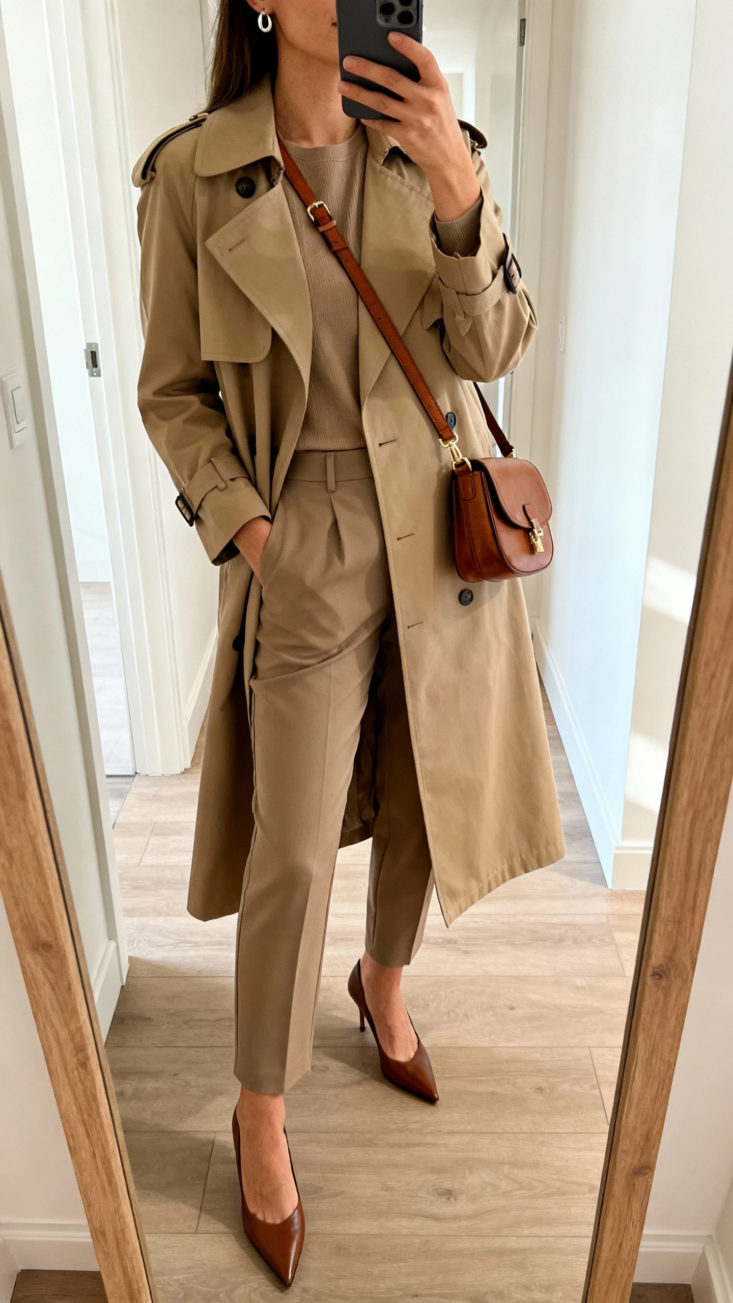 iPhone mirror selfie of a woman wearing a classic trench coat over a column of color (matching top and trousers), pointed pumps, leather crossbody, understated earrings, face not visible, cropped at shoulders, hallway mirror, natural daylight, iPhone photo quality.