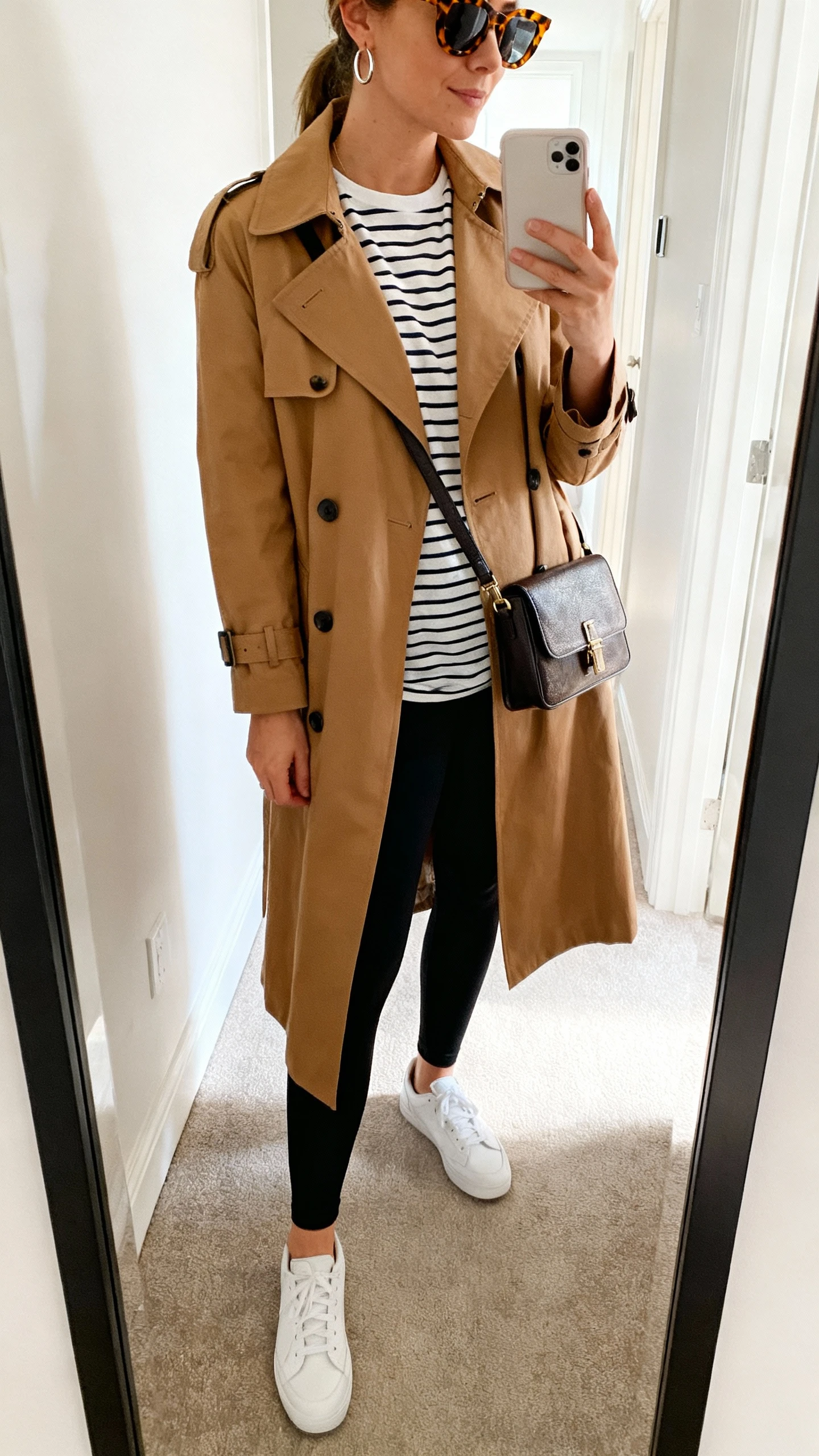iPhone mirror selfie of a woman wearing a classic camel trench coat over a striped Breton tee, matte black leggings, white sneakers, a structured crossbody, tortoiseshell sunglasses and delicate hoop earrings, face not visible, side angle in hallway mirror, bright morning light, iPhone photo quality.