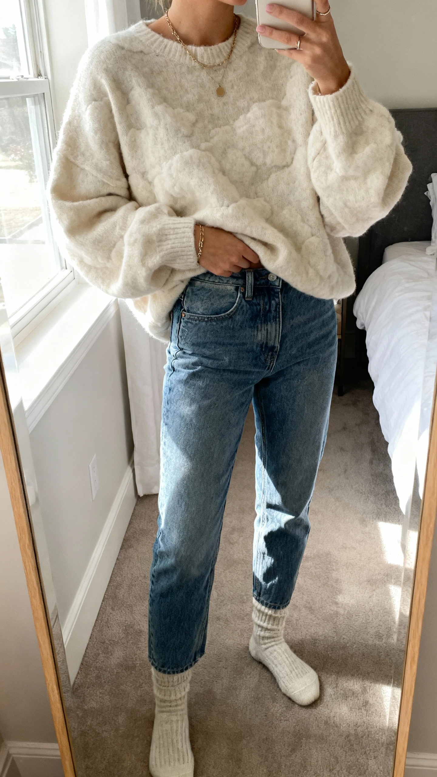 iPhone mirror selfie of a woman wearing a cloud-soft oversized sweater tucked into high-rise mom jeans with cozy socks and minimal jewelry, face not visible, cropped at neck, bedroom mirror, soft morning window light, iPhone photo quality.
