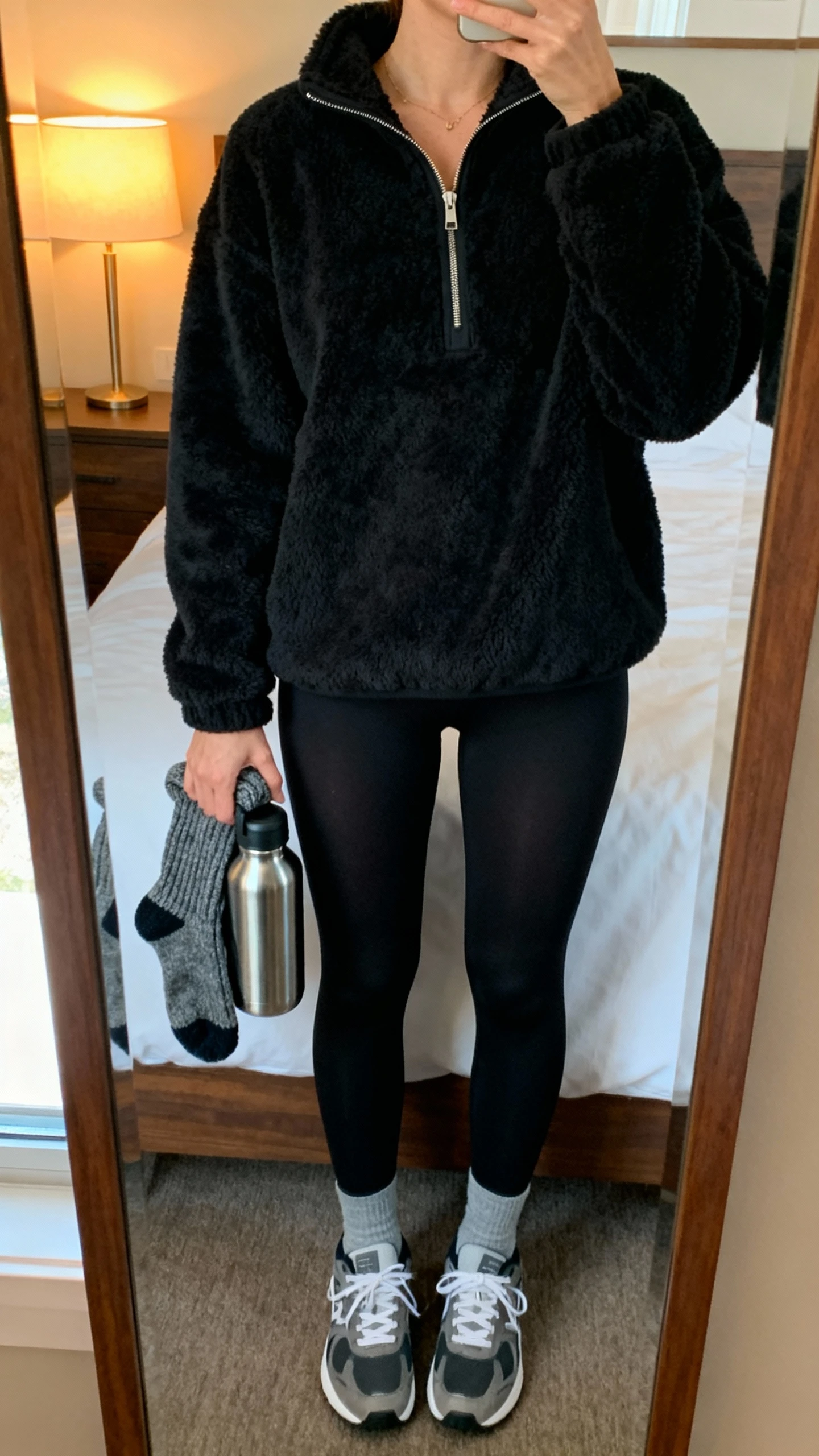 iPhone mirror selfie of a woman wearing a cozy black fleece half-zip and sleek black tights with cushy sneakers, face not visible, cropped at neck, bedroom mirror with warm lamp light, slouchy socks and a stainless water bottle in hand, iPhone photo quality.