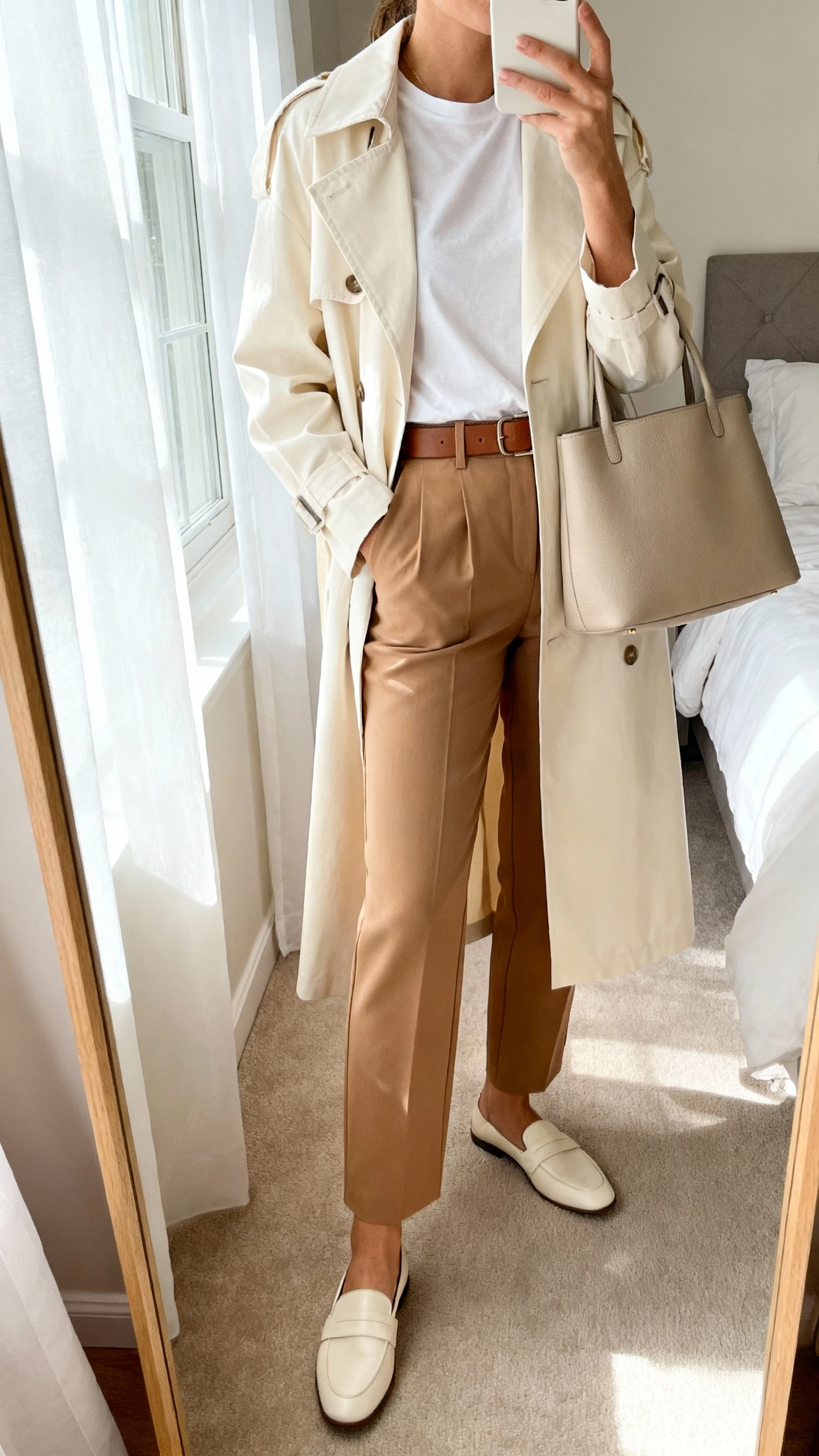 iPhone mirror selfie of a woman wearing a cream trench coat over a simple white tee, tailored tan trousers, minimalist leather belt, cream loafers, and a structured neutral tote, face not visible, cropped at neck, bedroom mirror, soft morning window light, iPhone photo quality.