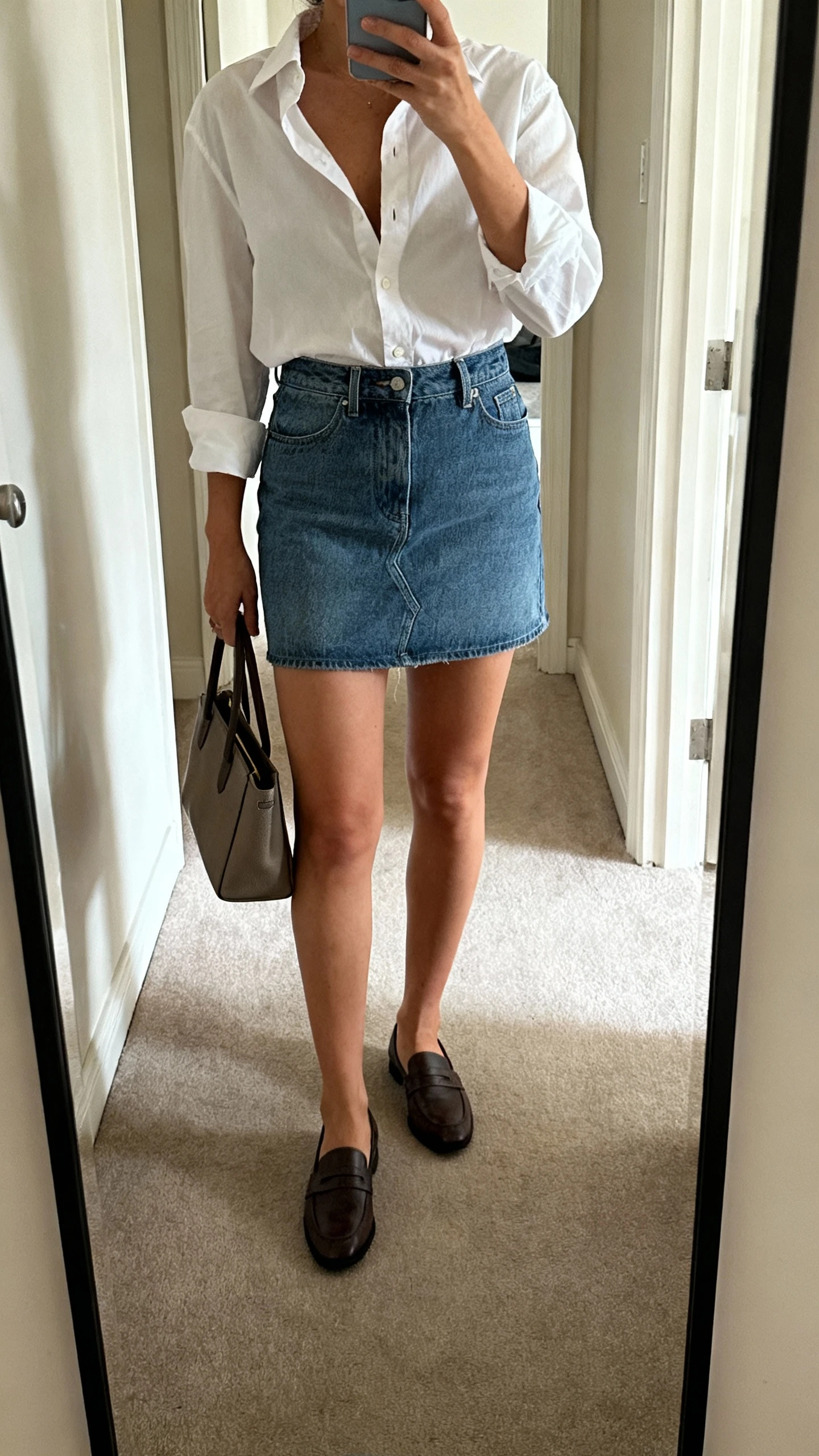 iPhone mirror selfie of a woman wearing a crisp button-up shirt (slightly unbuttoned) tucked into a denim mini skirt with loafers and a structured handbag, face not visible, cropped at shoulders, full-length hallway mirror, morning light, iPhone photo quality.