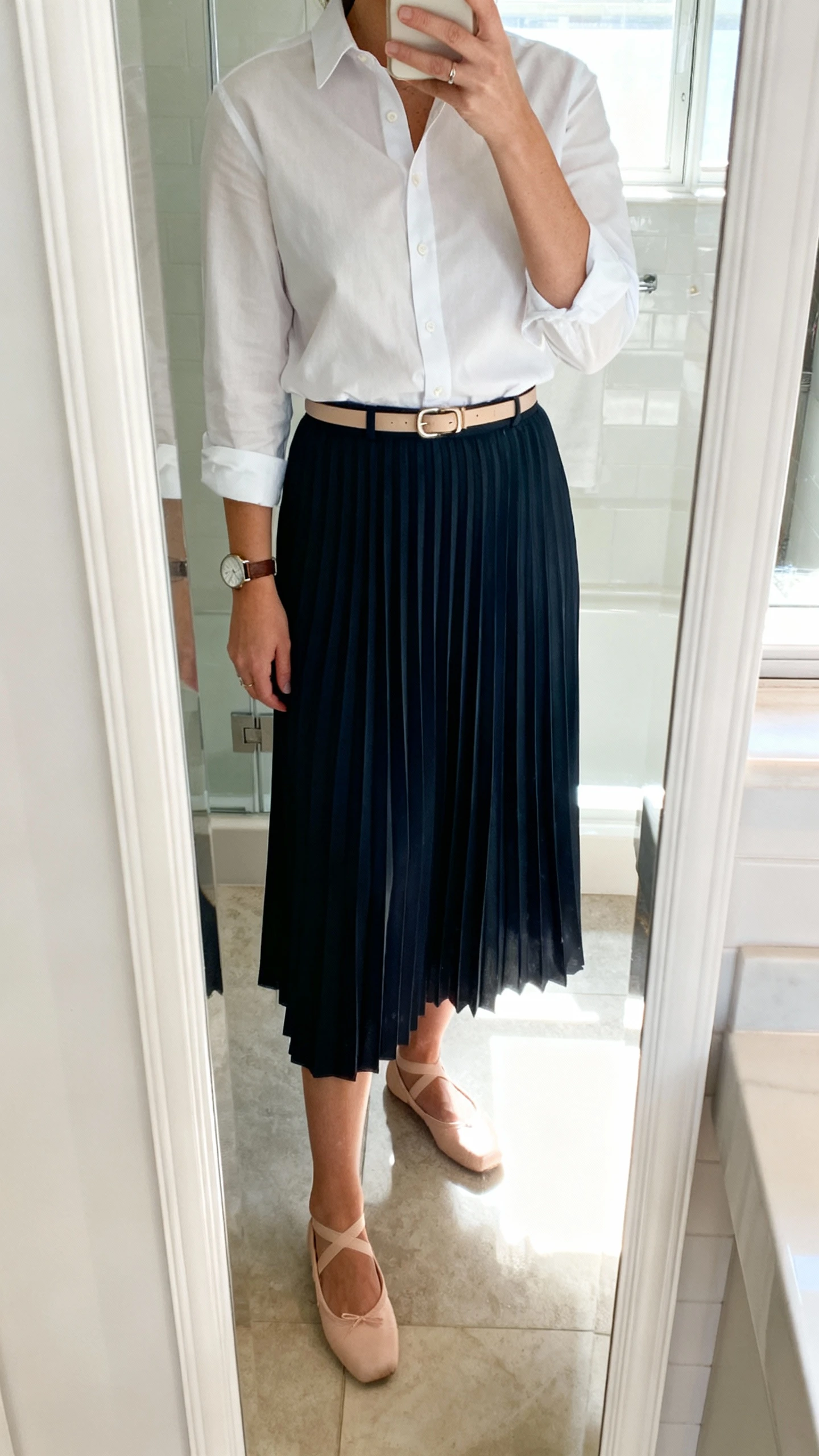 iPhone mirror selfie of a woman wearing a crisp button-down shirt tucked into a pleated midi skirt, ballet flats, slim belt, and a watch, face not visible, cropped at shoulders, bathroom mirror with bright indoor light, iPhone photo quality.