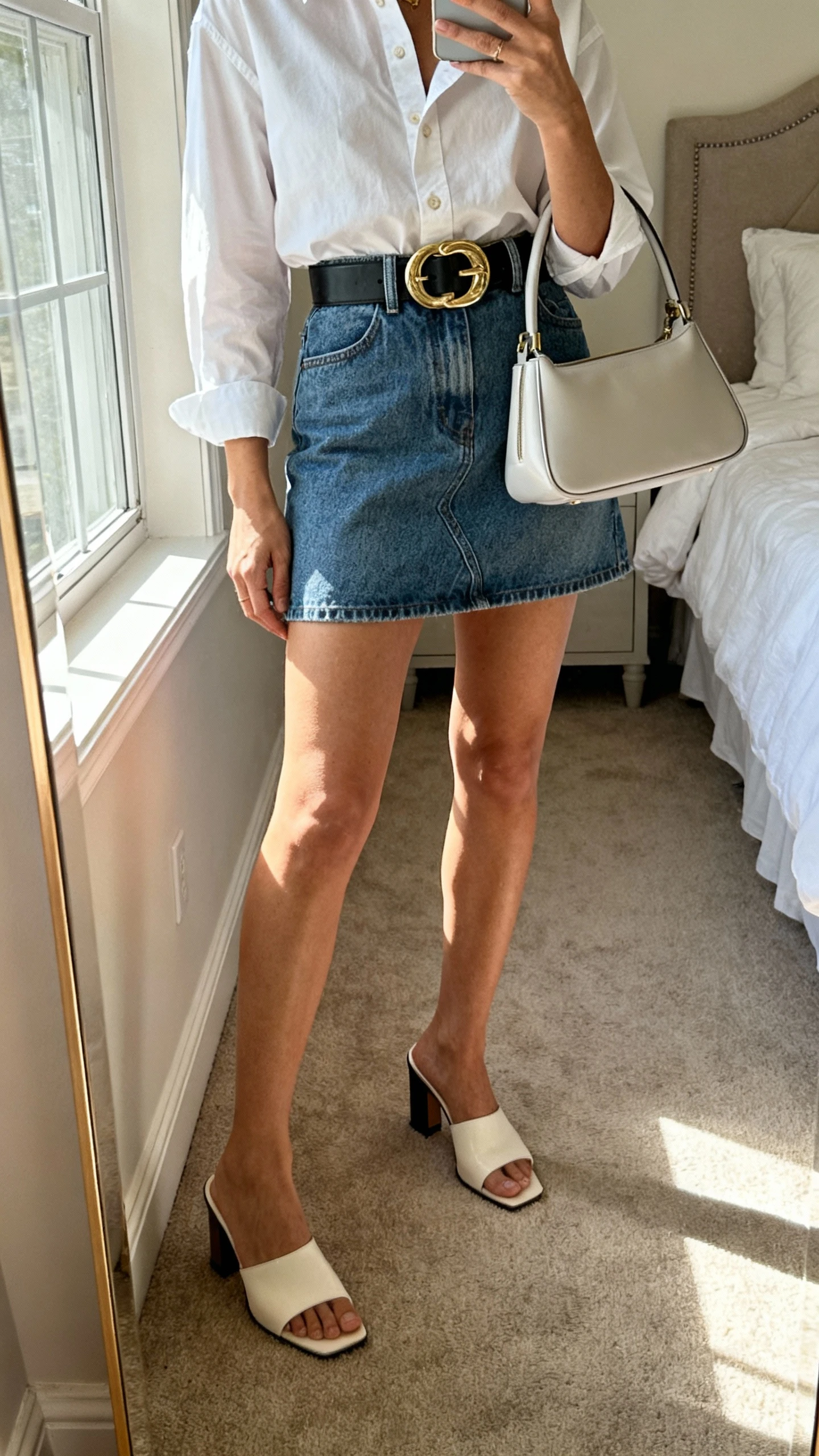 iPhone mirror selfie of a woman wearing a crisp button-down tucked into a denim mini skirt, wide statement belt, heeled mules, and a sleek top-handle bag, face not visible, bedroom mirror with morning window light, iPhone photo quality.