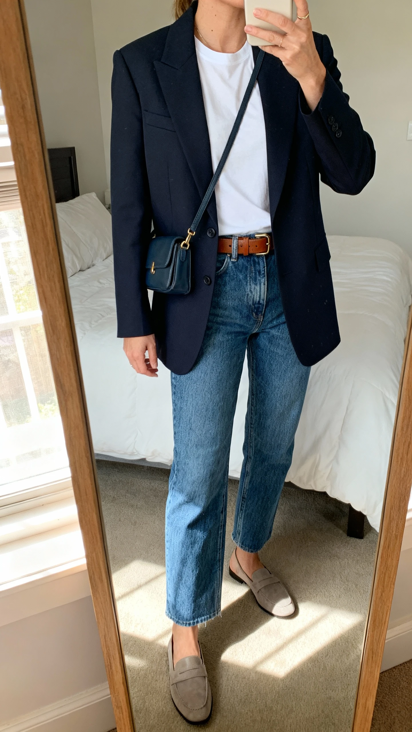 iPhone mirror selfie of a woman wearing a crisp structured blazer over a white tee, straight-leg blue jeans, leather belt, minimalist loafers, and a small crossbody bag, face not visible, cropped at neck, bedroom mirror with natural window light, iPhone photo quality.