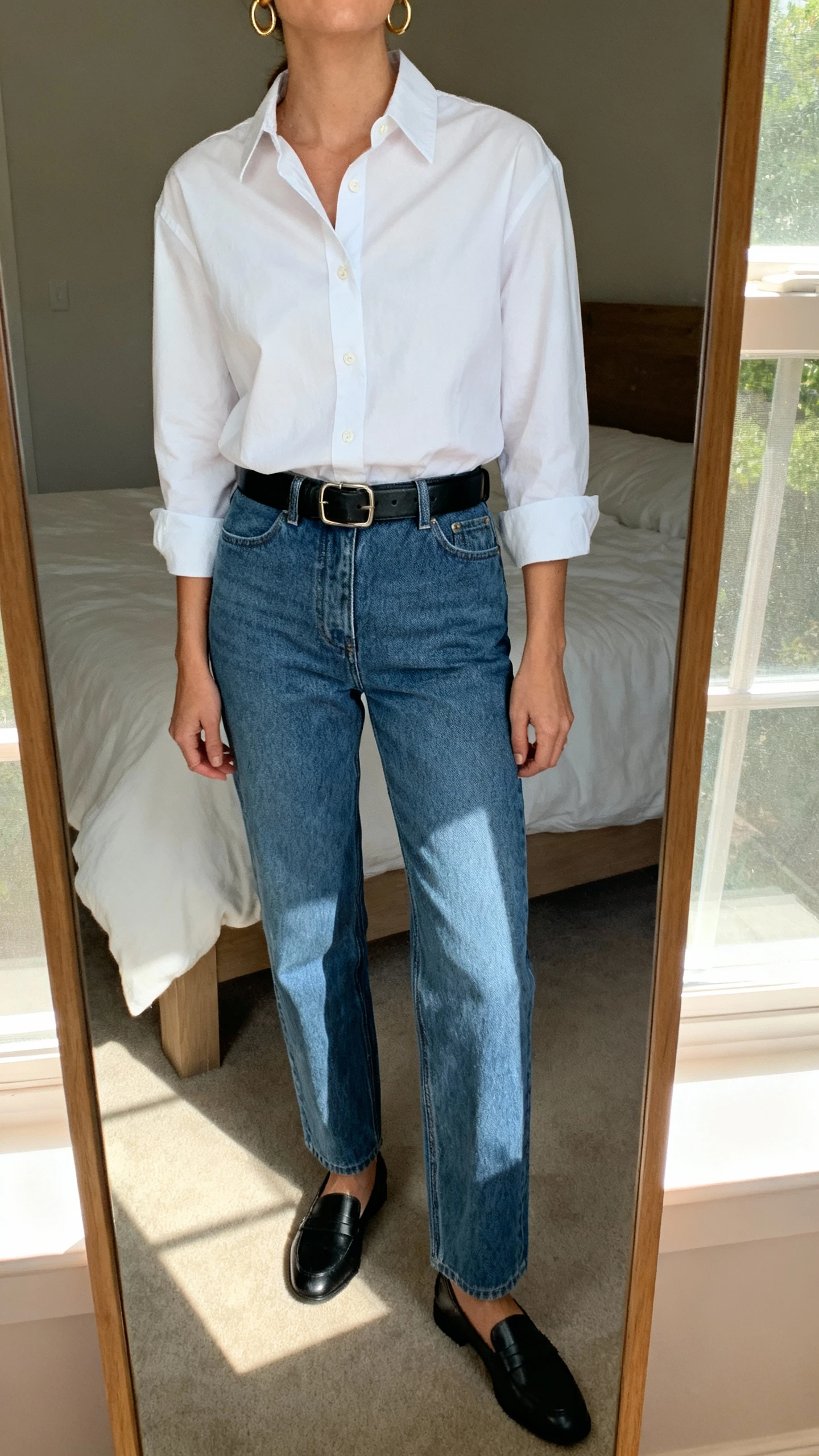 iPhone mirror selfie of a woman wearing a crisp white button-down tucked into straight-leg blue jeans, black leather belt, minimalist gold hoops, black loafers, face not visible, cropped at neck, bedroom mirror with natural window light, iPhone photo quality