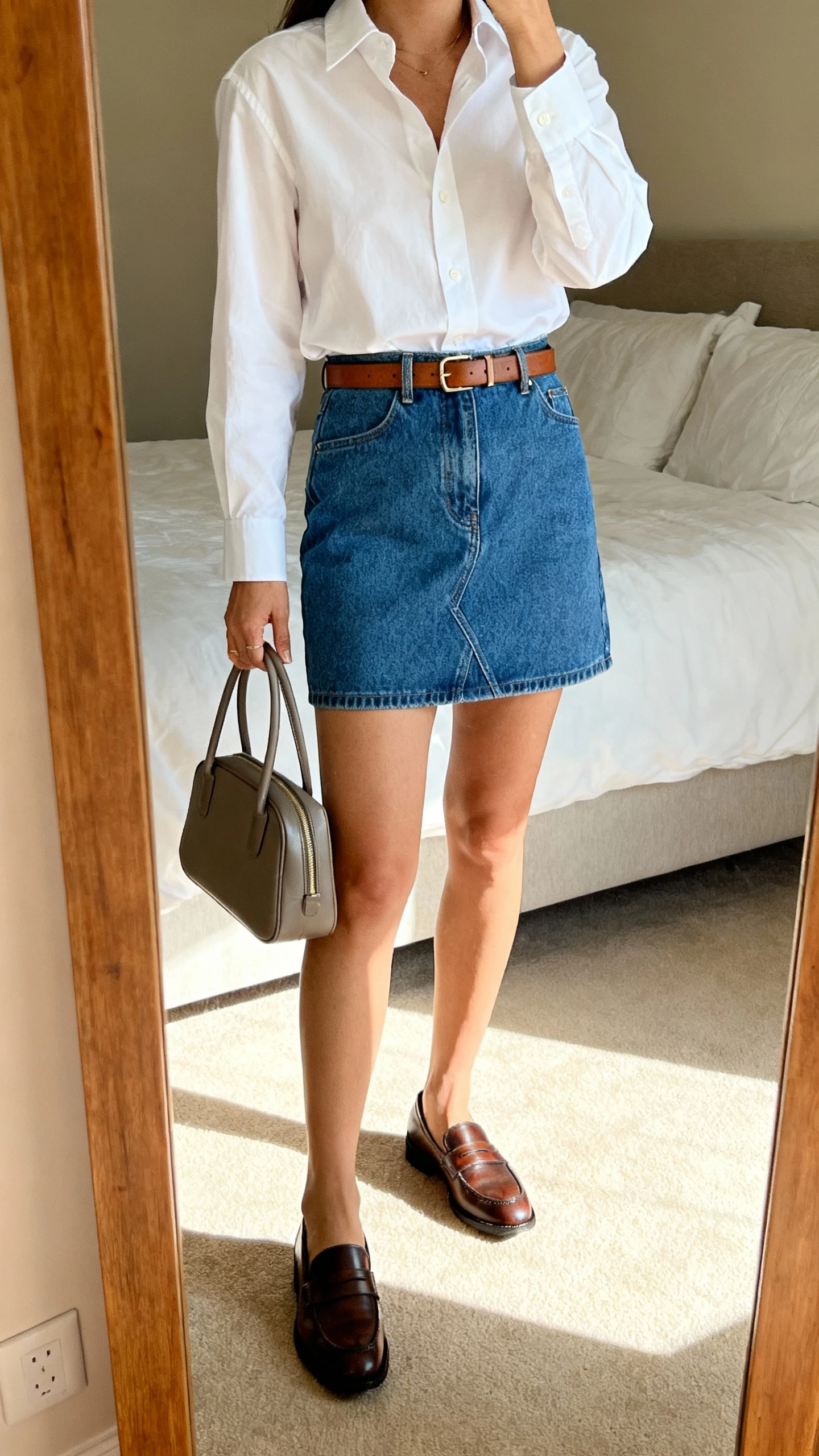 iPhone mirror selfie of a woman wearing a crisp white button-down shirt tucked into a blue denim mini skirt, penny loafers, and a slim leather belt with a structured top-handle bag, face not visible, cropped at shoulders, bedroom mirror, morning light, iPhone photo quality.