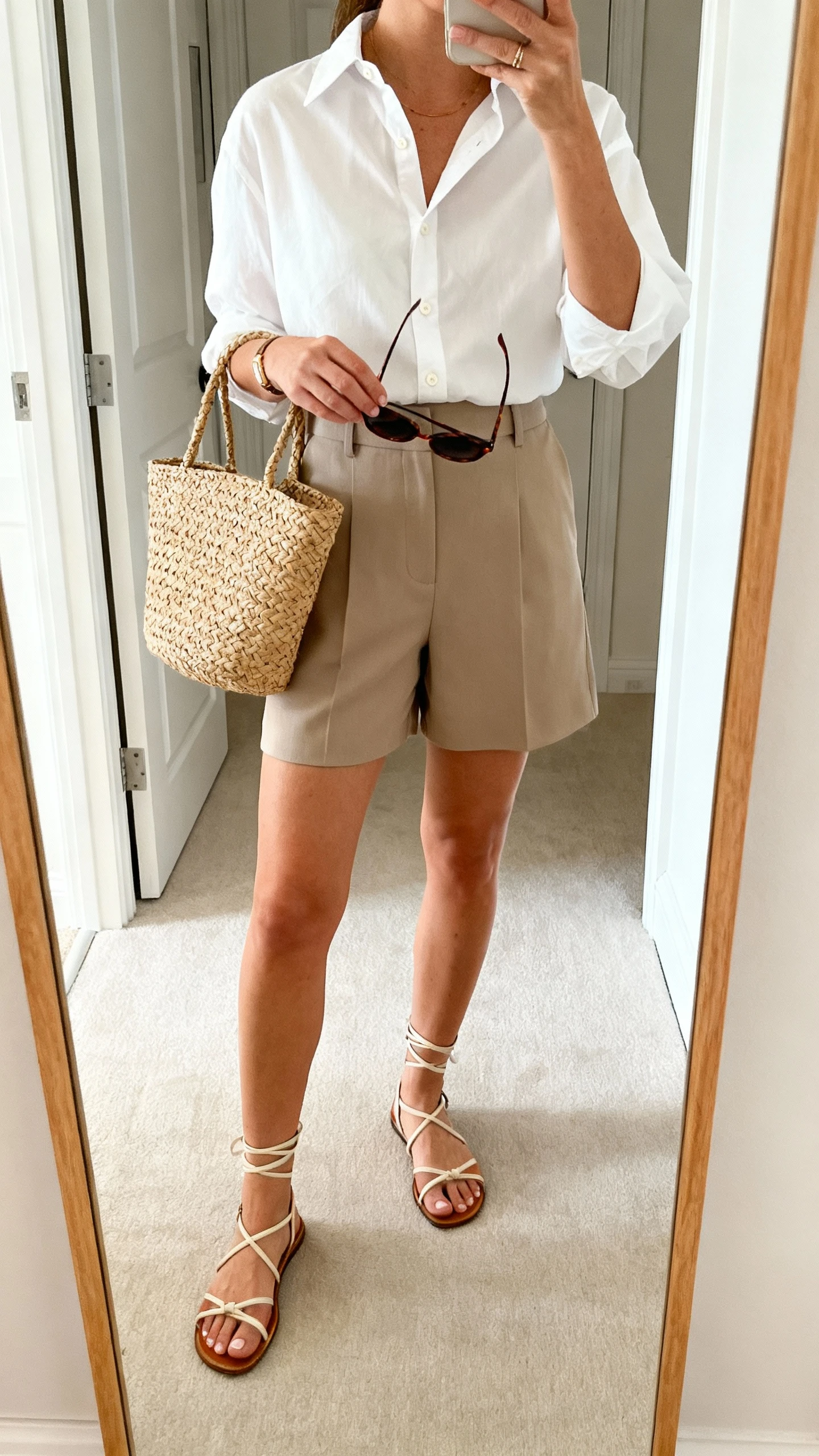 iPhone mirror selfie of a woman wearing a crisp white button-up tucked into tailored high-waist shorts, strappy sandals, a woven tote, and sunglasses in hand, face not visible, cropped at shoulders, entryway mirror, bright natural light, iPhone photo quality.