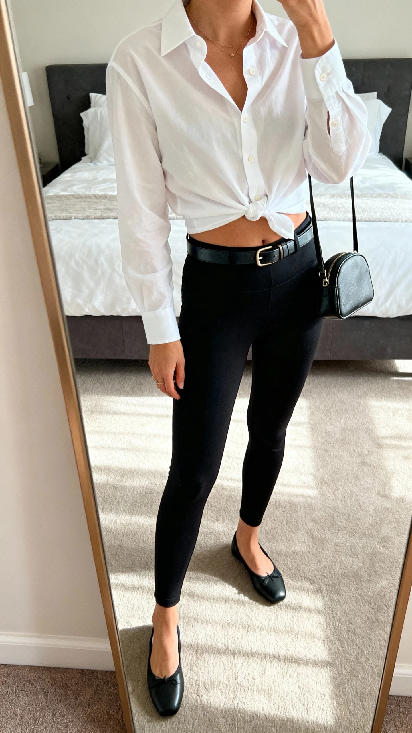 iPhone mirror selfie of a woman wearing a crisp white button-up shirt half-tucked into belted black leggings, slim leather belt with small buckle, ballet flats, structured mini bag, face not visible, cropped at neck, bedroom mirror, soft daylight, iPhone photo quality.
