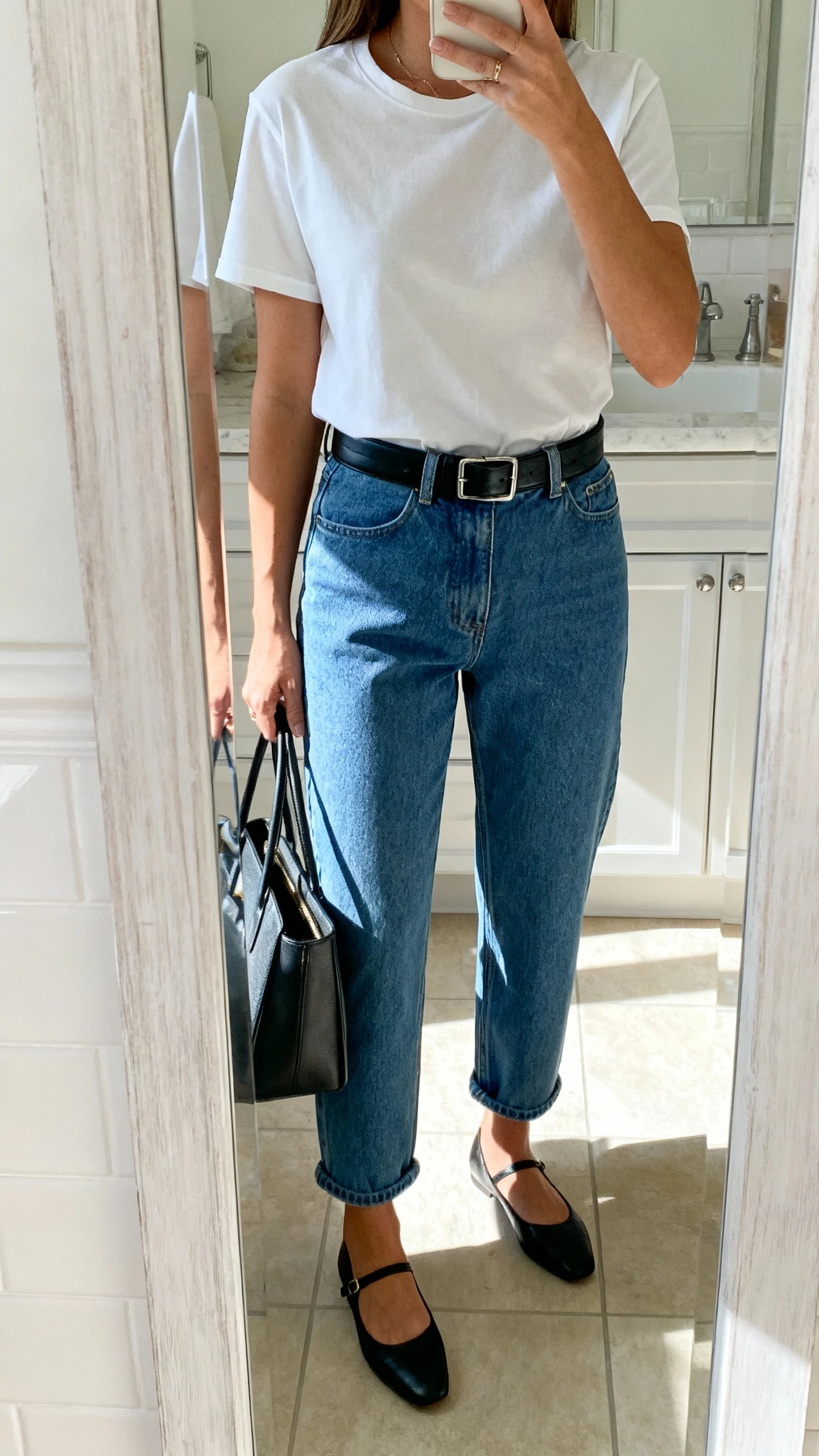 iPhone mirror selfie of a woman wearing a crisp white tee tucked into barrel-leg blue jeans, black leather belt, chic black ballet flats, structured tote, face not visible, shoulder-cropped, bathroom mirror, bright daylight, iPhone photo quality.