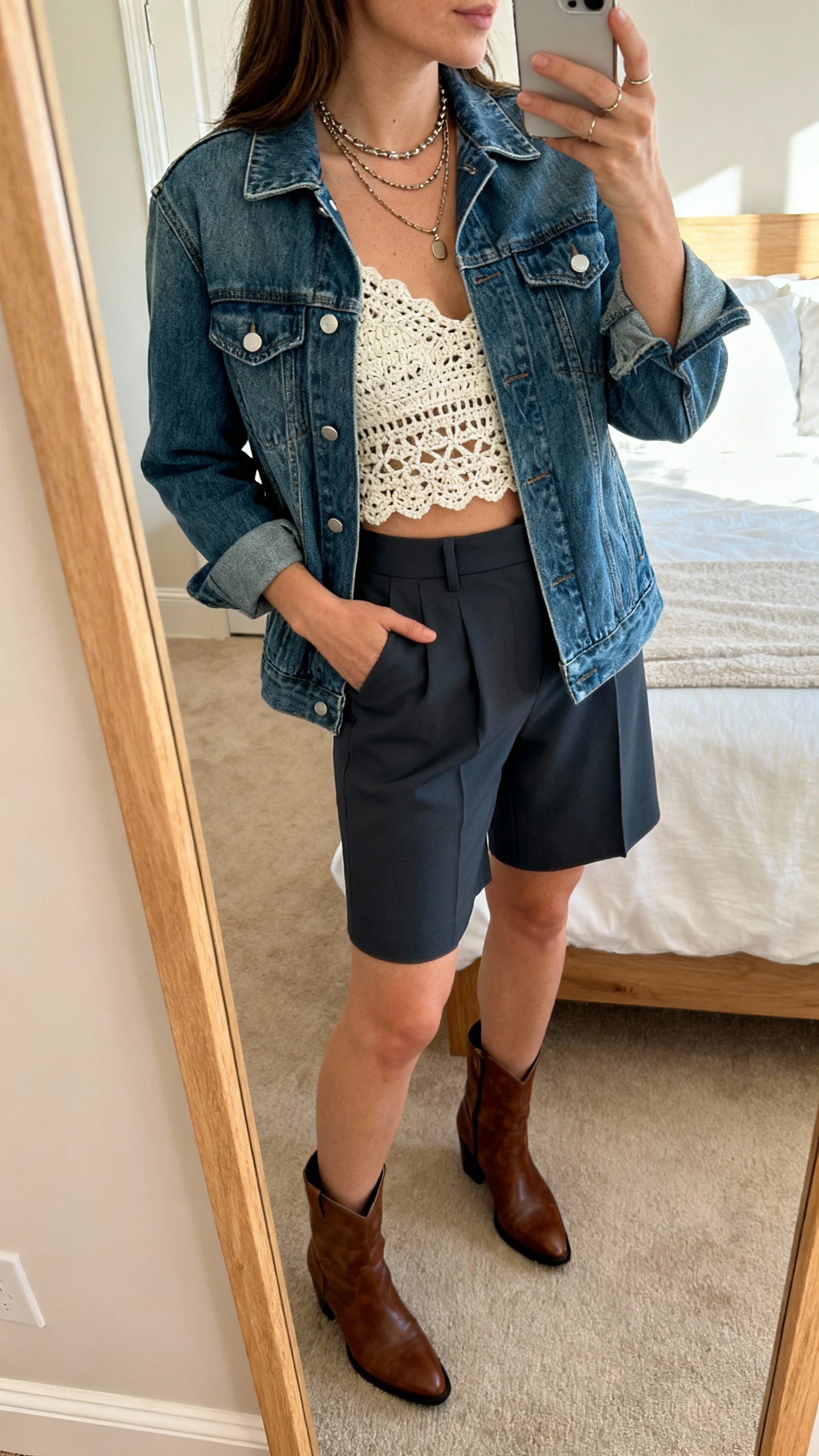 iPhone mirror selfie of a woman wearing a denim jacket over a crochet top with tailored shorts, ankle boots, and layered necklaces, face not visible, side angle, bedroom mirror, afternoon natural light, iPhone photo quality.
