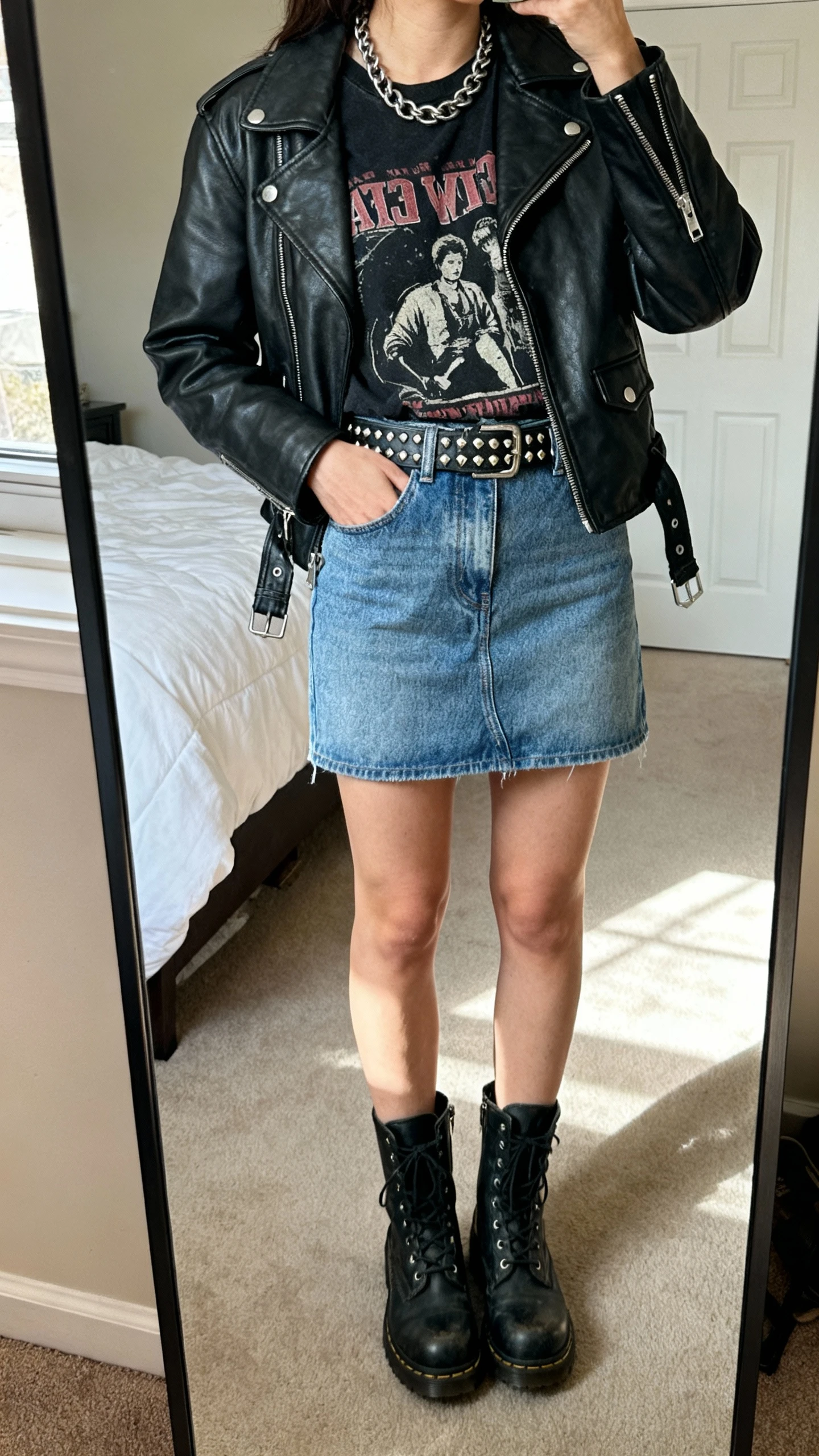iPhone mirror selfie of a woman wearing a denim mini skirt, vintage black graphic tee tucked in, leather moto jacket, combat boots, chain necklace, and a studded belt, face not visible, cropped at shoulders, bedroom mirror, natural daylight, iPhone photo quality.
