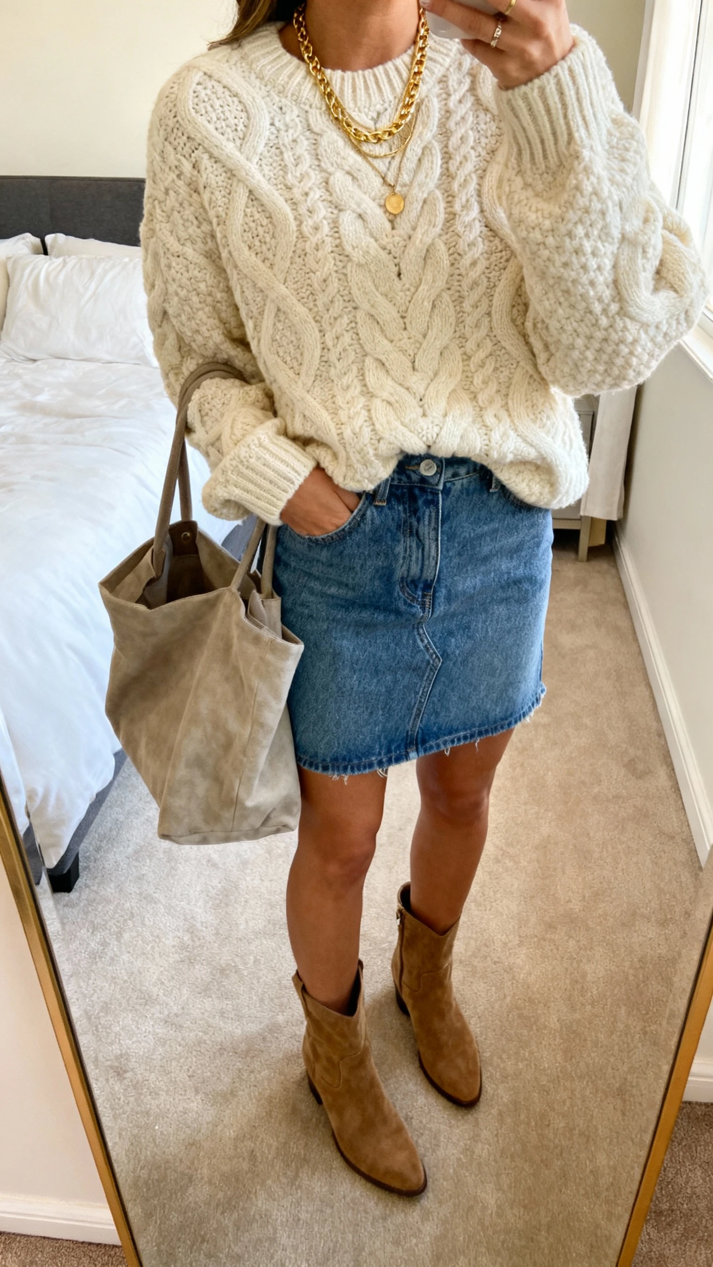 iPhone mirror selfie of a woman wearing a denim mini skirt, chunky cream cable-knit sweater, suede ankle boots, slouchy tote, and layered gold necklaces, face not visible, cropped at shoulders, bedroom mirror, warm indoor lighting, iPhone photo quality.