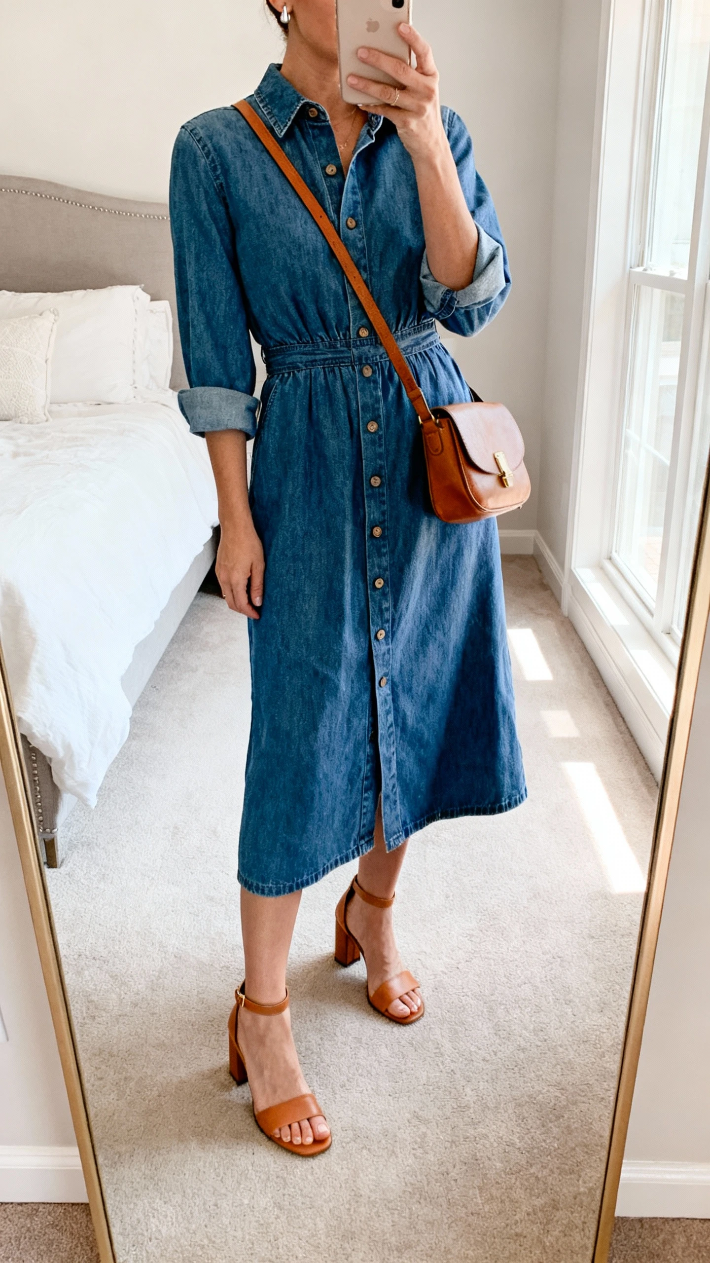 iPhone mirror selfie of a woman wearing a denim midi dress with a defined waist, cuffed sleeves, and front buttons, tan heeled sandals, leather crossbody, simple studs, face not visible, cropped at neck, bedroom mirror, natural light, iPhone photo quality.