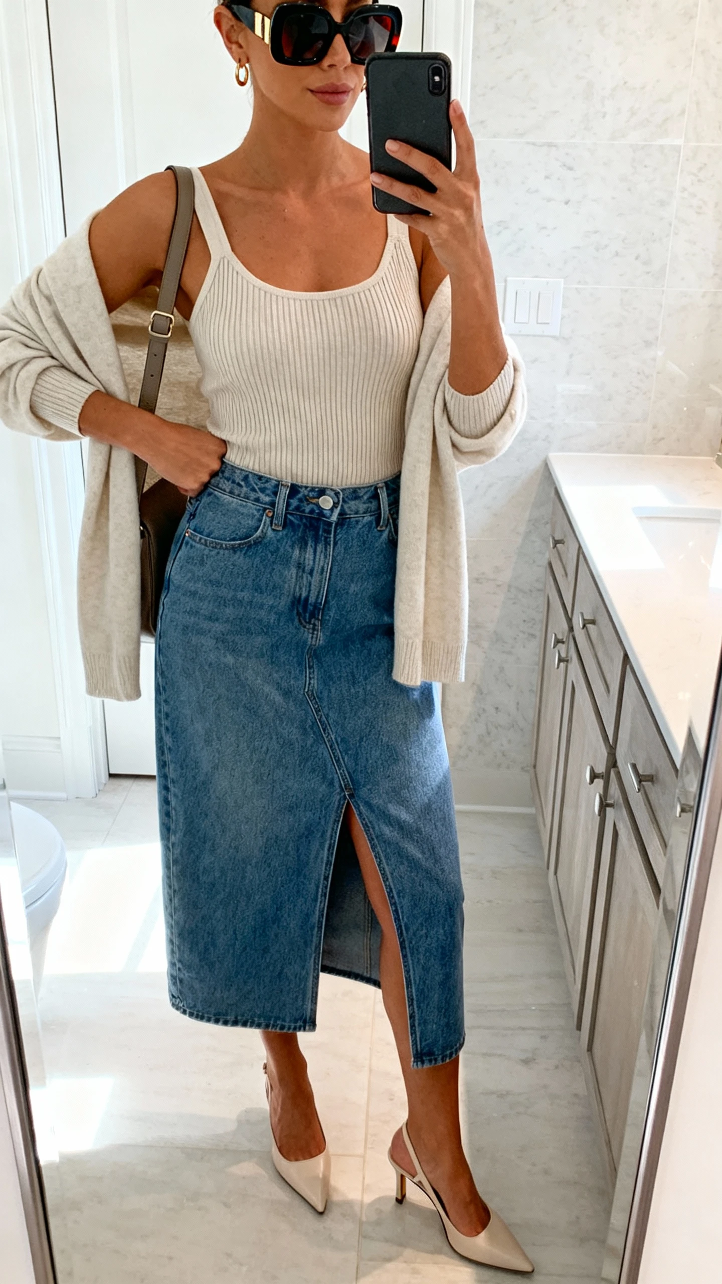 iPhone mirror selfie of a woman wearing a denim midi skirt with front slit, ribbed knit tank, kitten-heel slingbacks, lightweight cardigan over shoulders, shoulder bag, bold sunglasses, and simple earrings; face not visible, bathroom mirror, bright indoor lighting, iPhone photo quality.