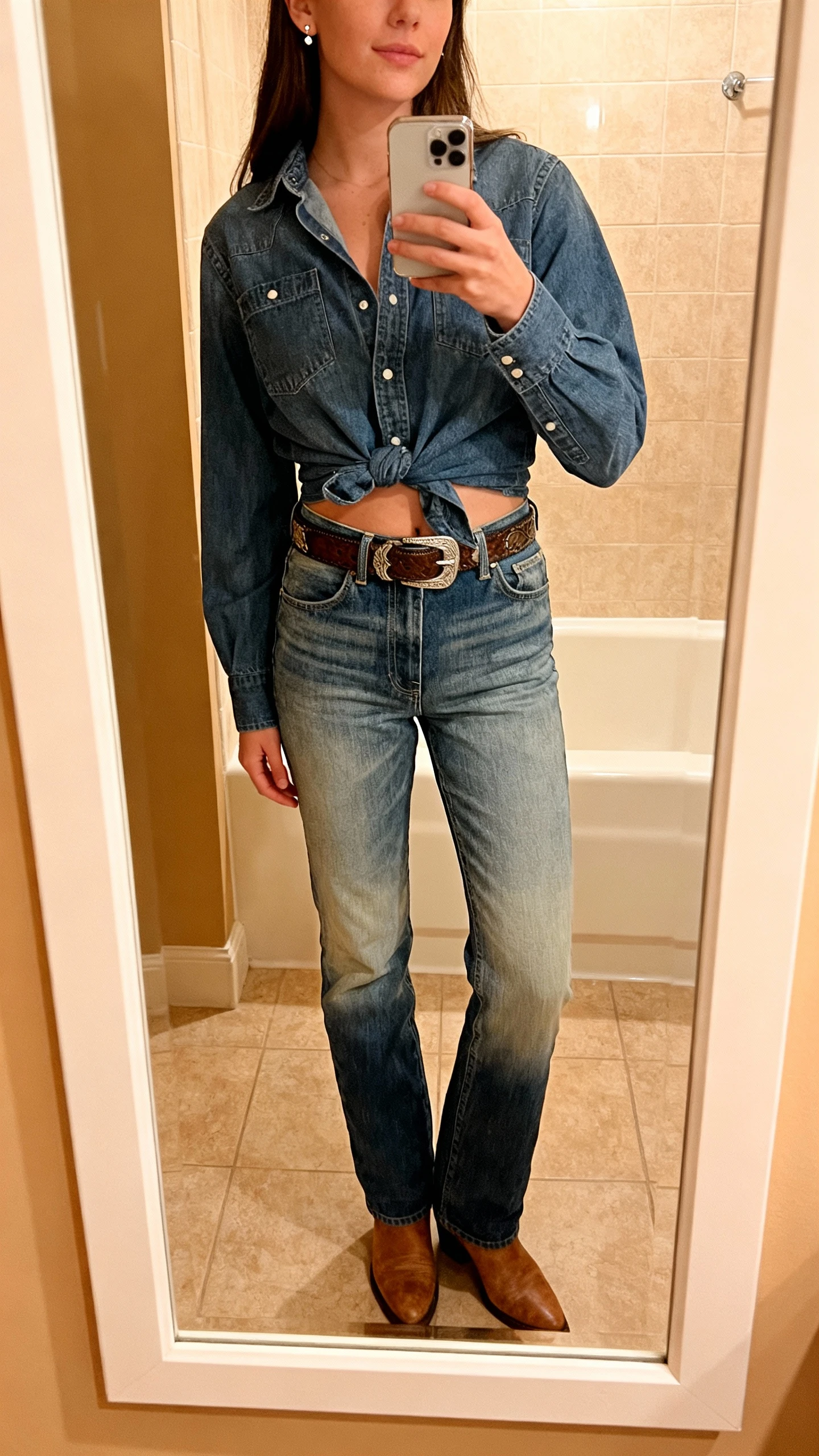 iPhone mirror selfie of a woman wearing a denim shirt tucked into high-rise denim jeans (slightly different wash), Western belt, ankle boots, and simple stud earrings, face not visible, bathroom mirror, warm indoor light, iPhone photo quality.