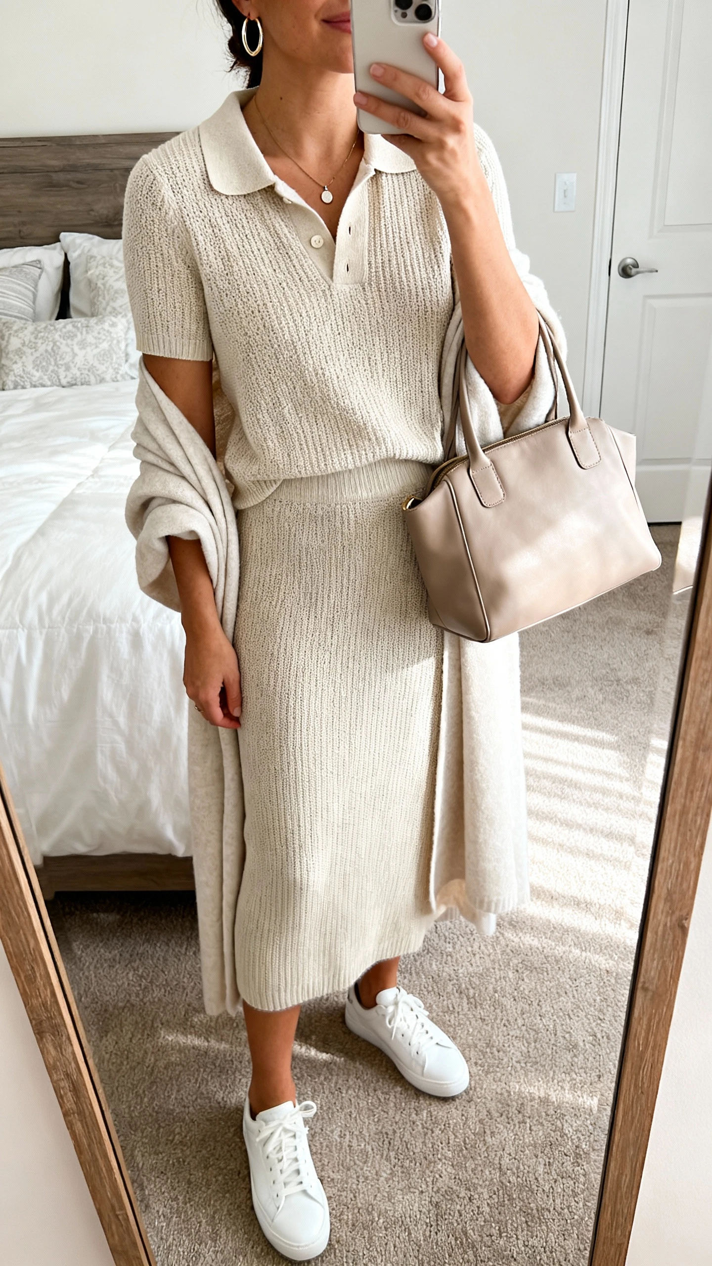 iPhone mirror selfie of a woman wearing a fine knit polo with matching midi knit skirt, lightweight duster, clean white sneakers, minimal pendant, hoop earrings, and a soft leather tote; face not visible, cropped at neck, bedroom mirror with soft morning light, iPhone photo quality.