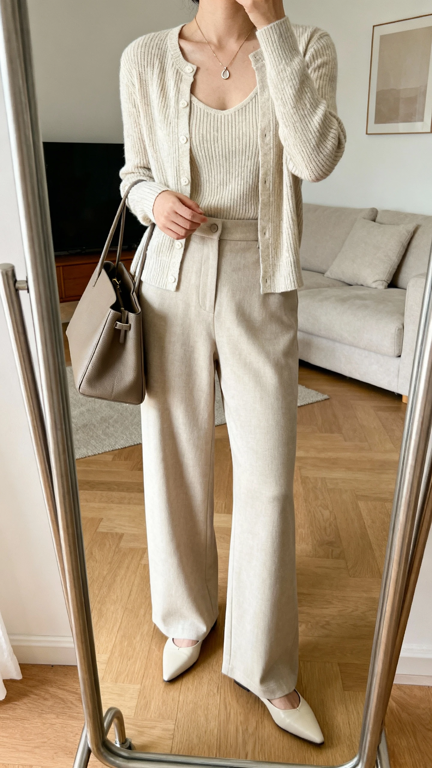 iPhone mirror selfie of a woman wearing a fine-knit cardigan set (shell + matching cardigan) tucked into high-waist wide-leg trousers, pointed flats, structured tote, and a delicate pendant necklace, face not visible, cropped at neck, living room mirror, soft indoor lighting, iPhone photo quality